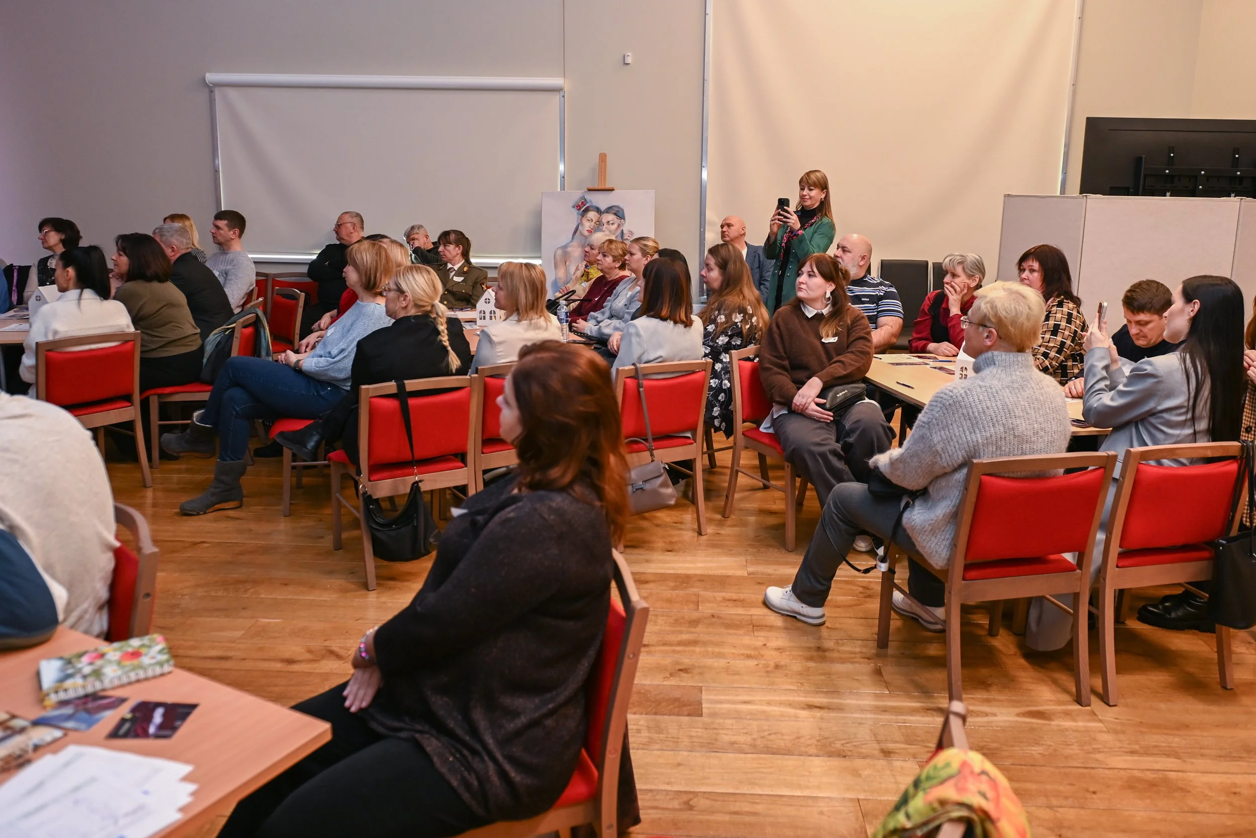 People attending a conference or seminar in a room with red chairs, a wooden floor, and a large white projection screen, one woman standing and taking a photo with her phone.