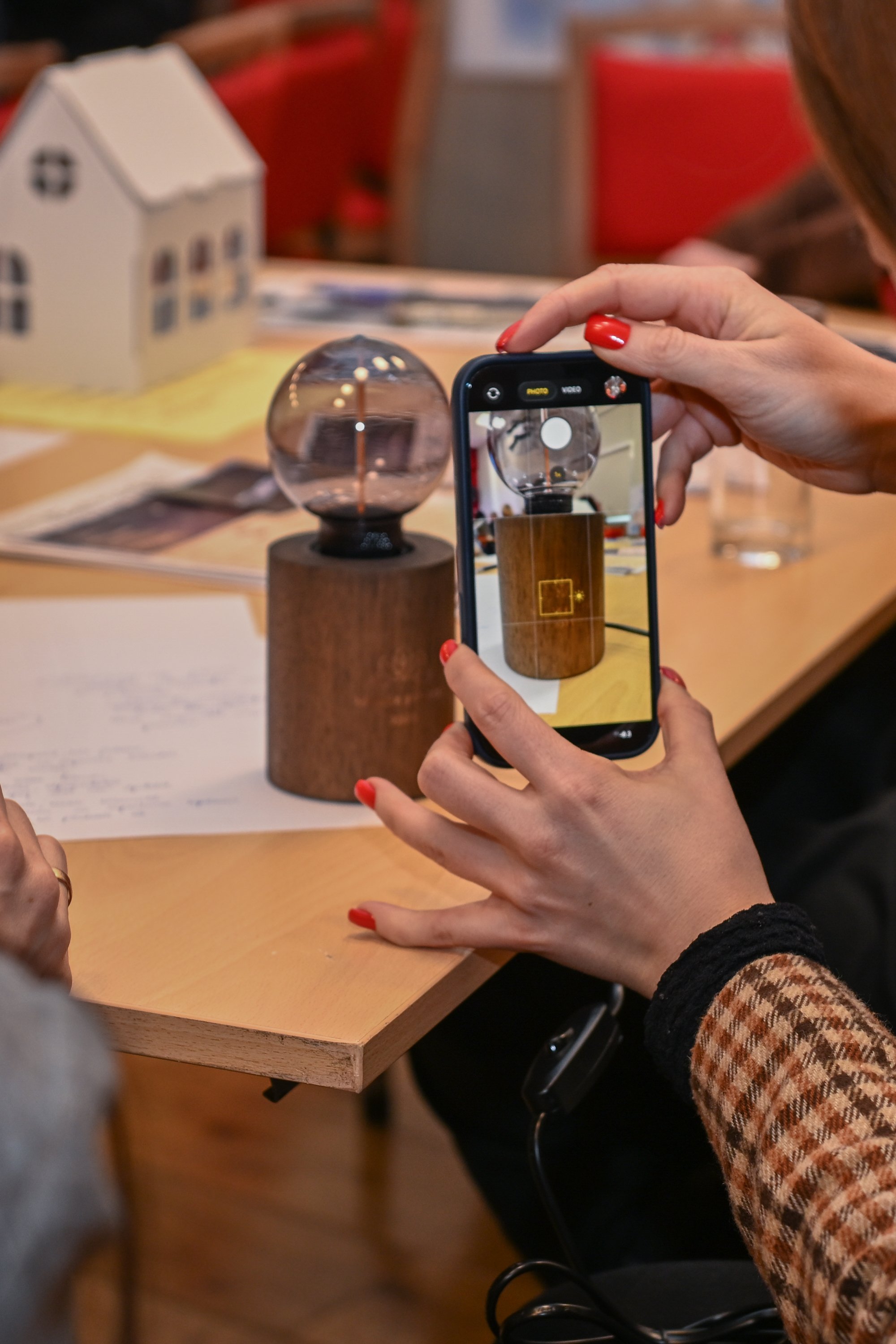 A person is taking a photo of a small spherical glass object on a wooden base using a smartphone, with another person partially visible nearby. The setting appears to be an indoor space with a table, papers, and a model house in the background.
