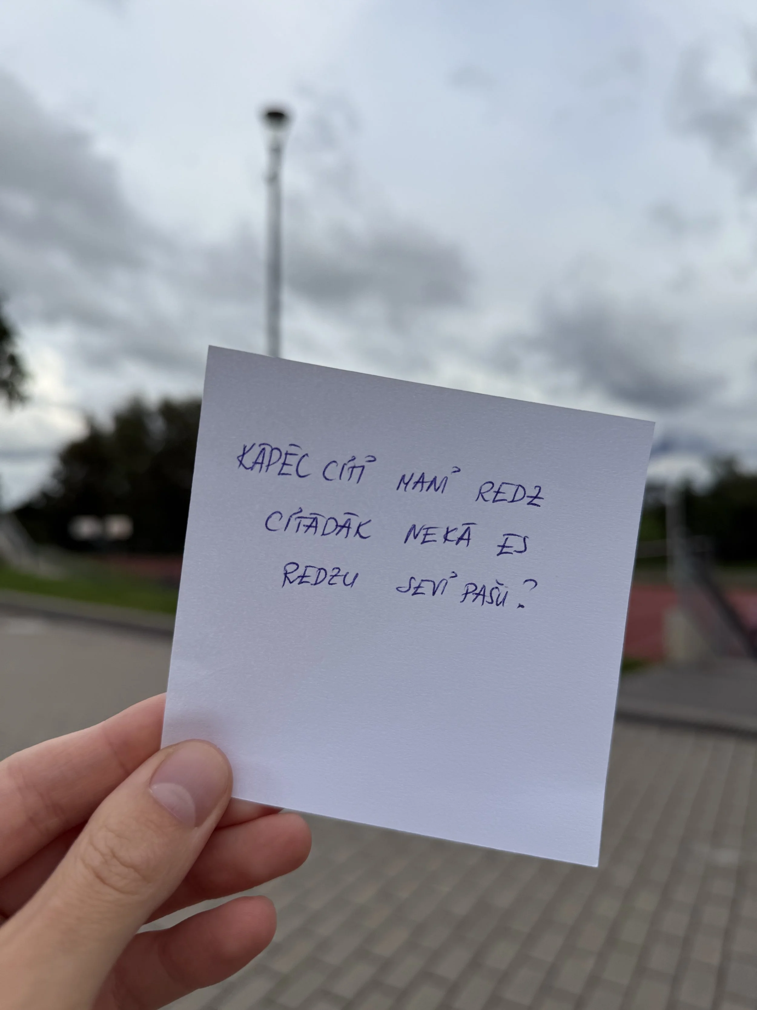 Hand holding a paper with handwritten text in Slovak, outdoors with cloudy sky, streetlamp, trees, and pavement in the background.