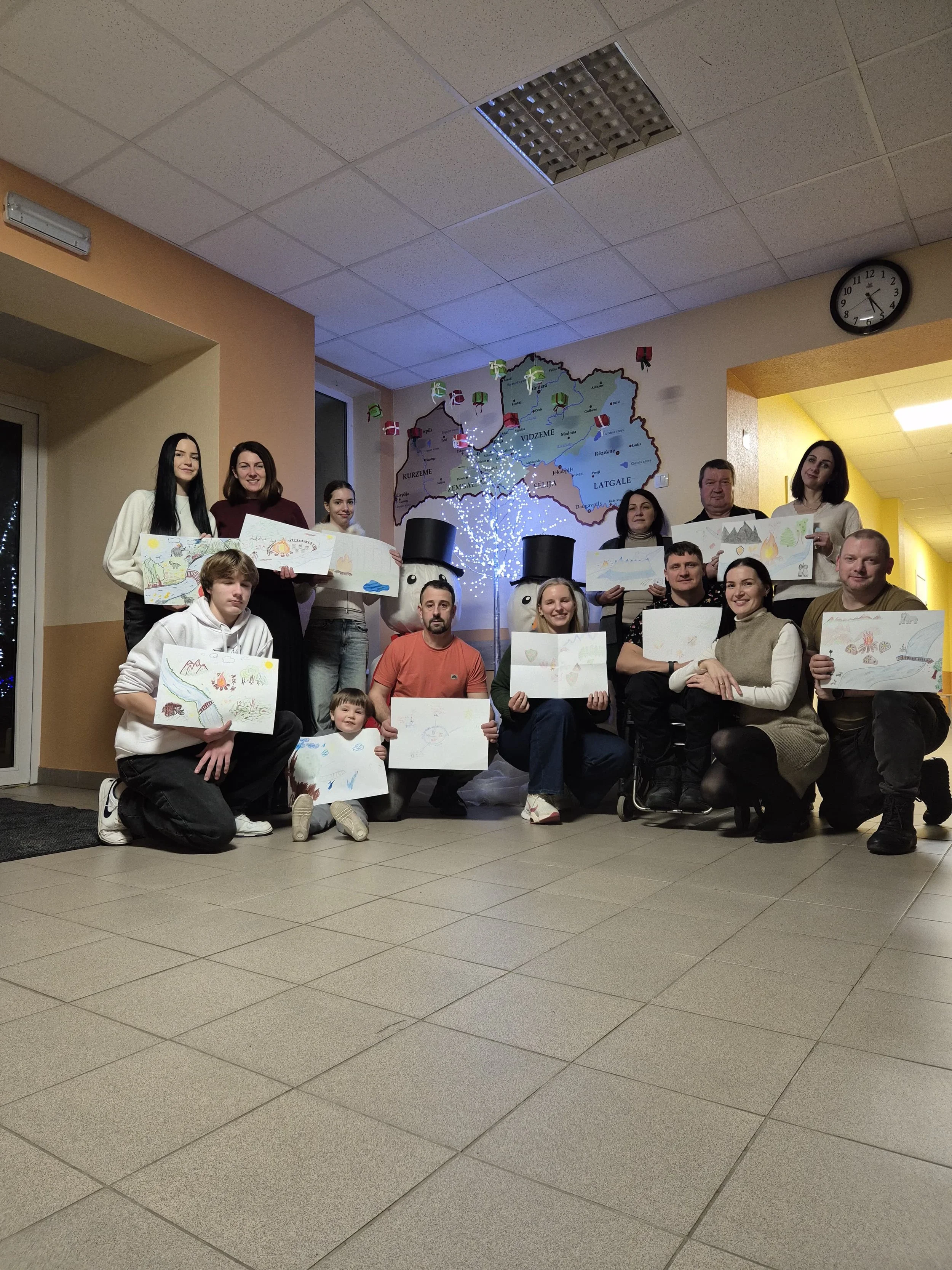 Group of people holding drawings in an indoor setting with a large map and decorative figures of snowmen in the background.