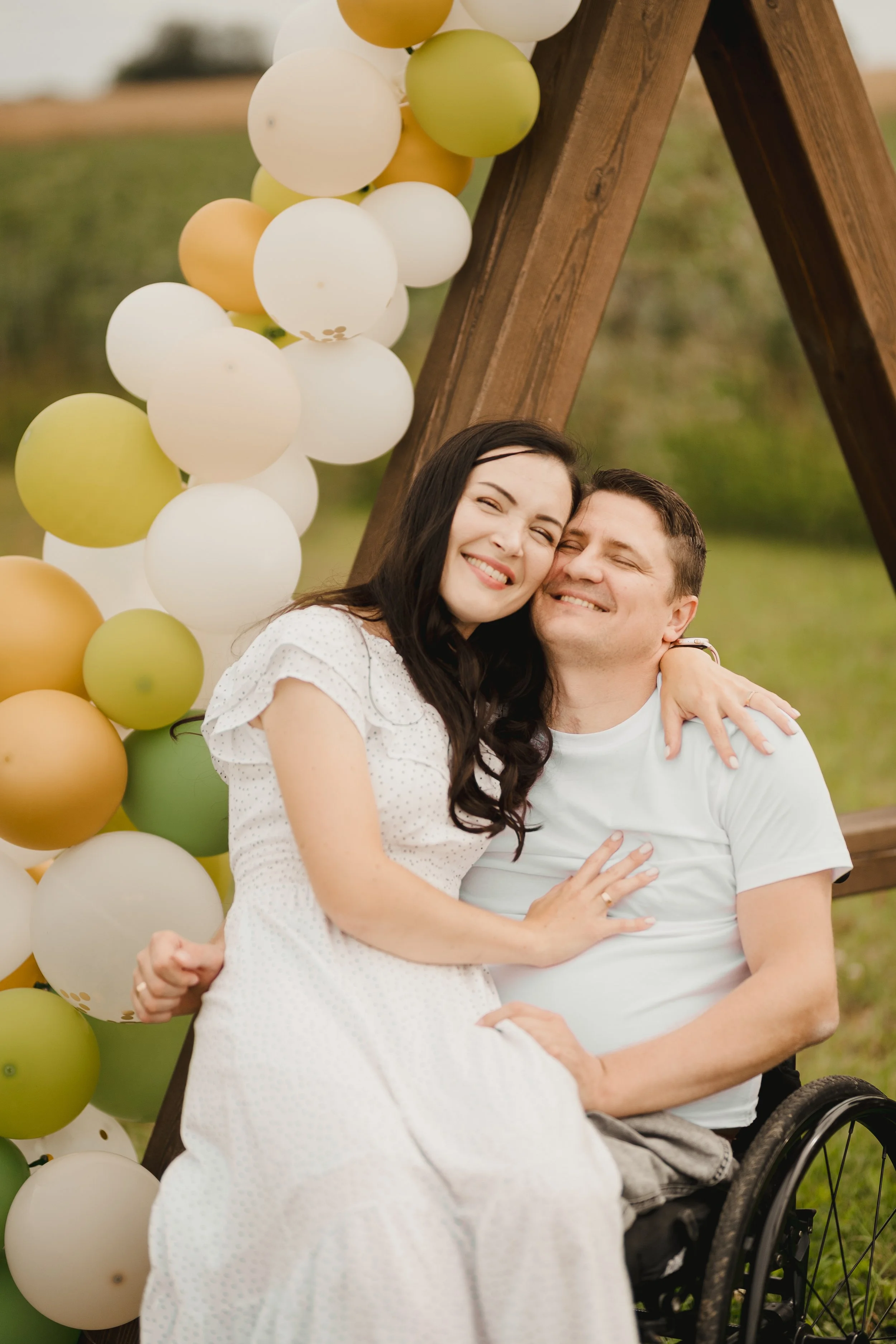 A happy woman and man in a wheelchair hugging each other outdoors near a wooden structure decorated with white, light green, and tan balloons.