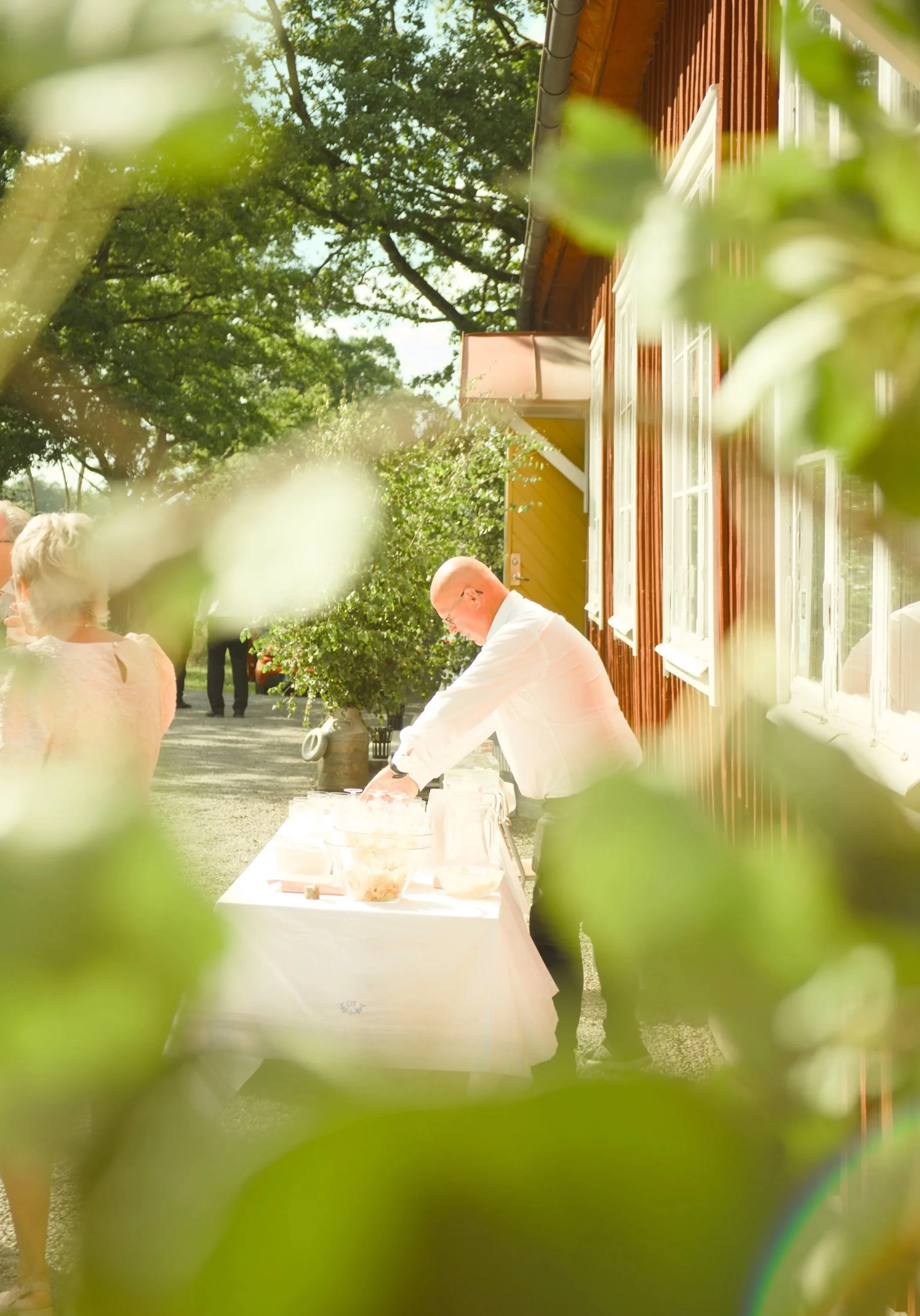 A man wearing a white shirt and black pants preparing food or drinks at a table outdoors during daytime, framed by green leaves with a red building and trees in the background.