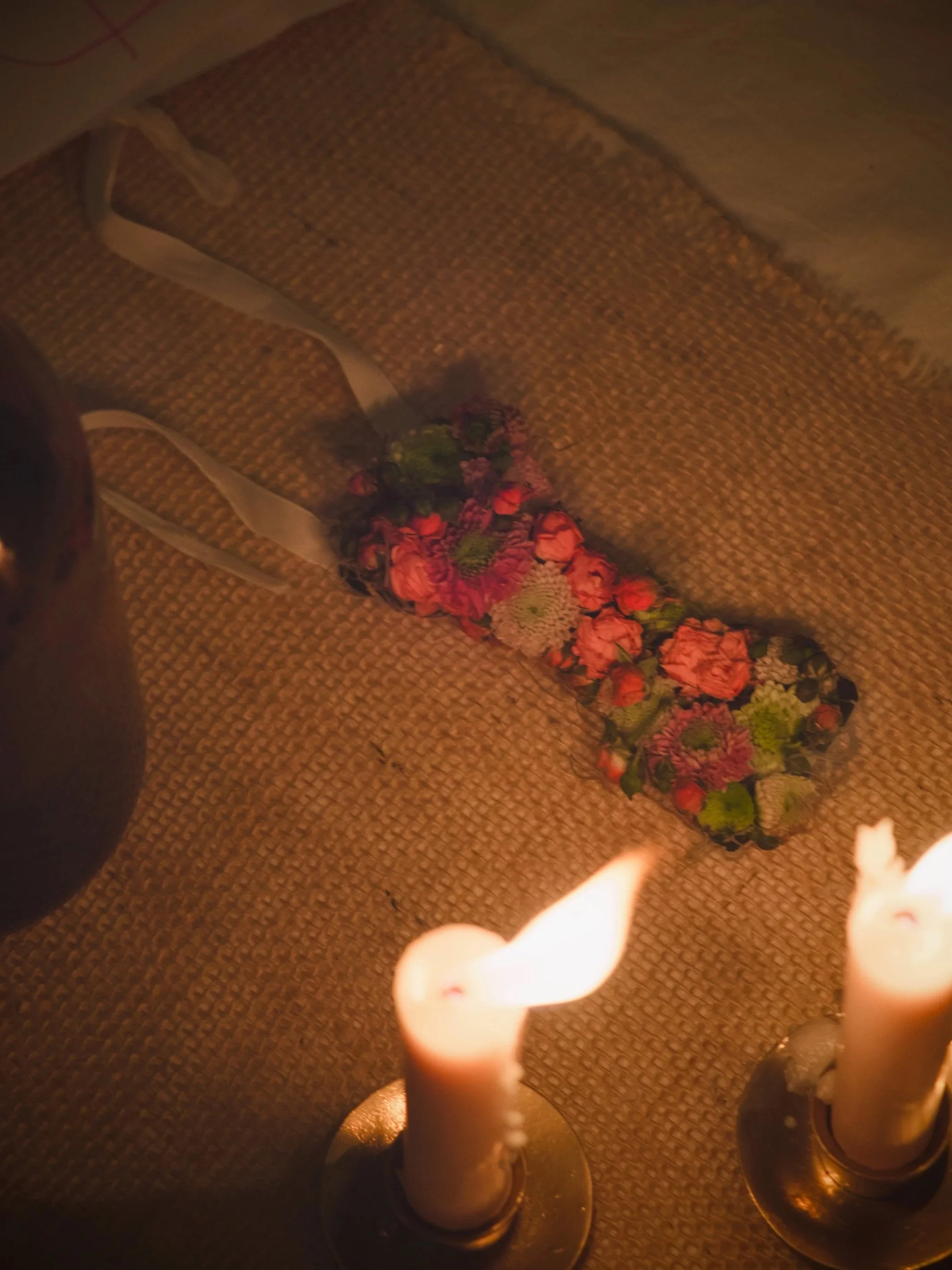 Two lit candles on a surface, with a floral headband or bracelet made of pink, red, and white flowers placed nearby.