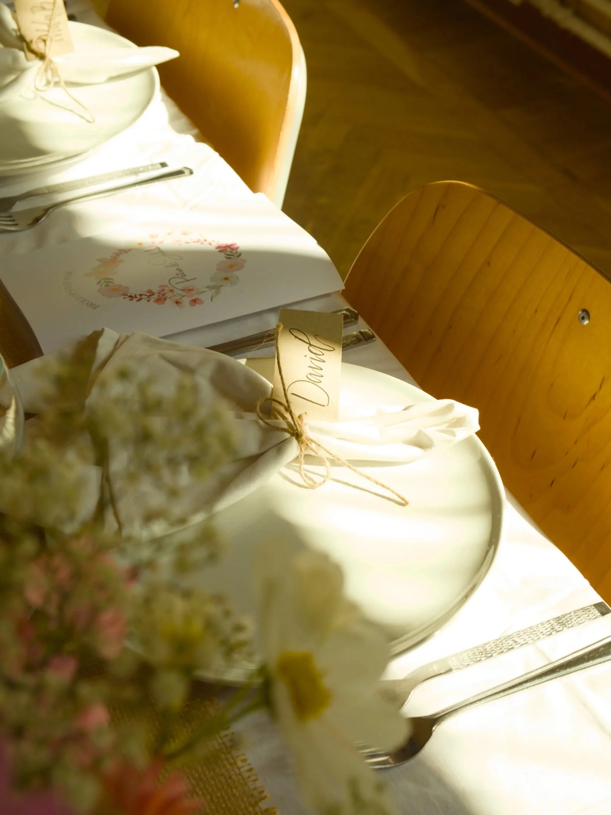 A set dining table with white tablecloth, cutlery, a place card labeled 'David', a menu with floral design, and a small bouquet of flowers.
