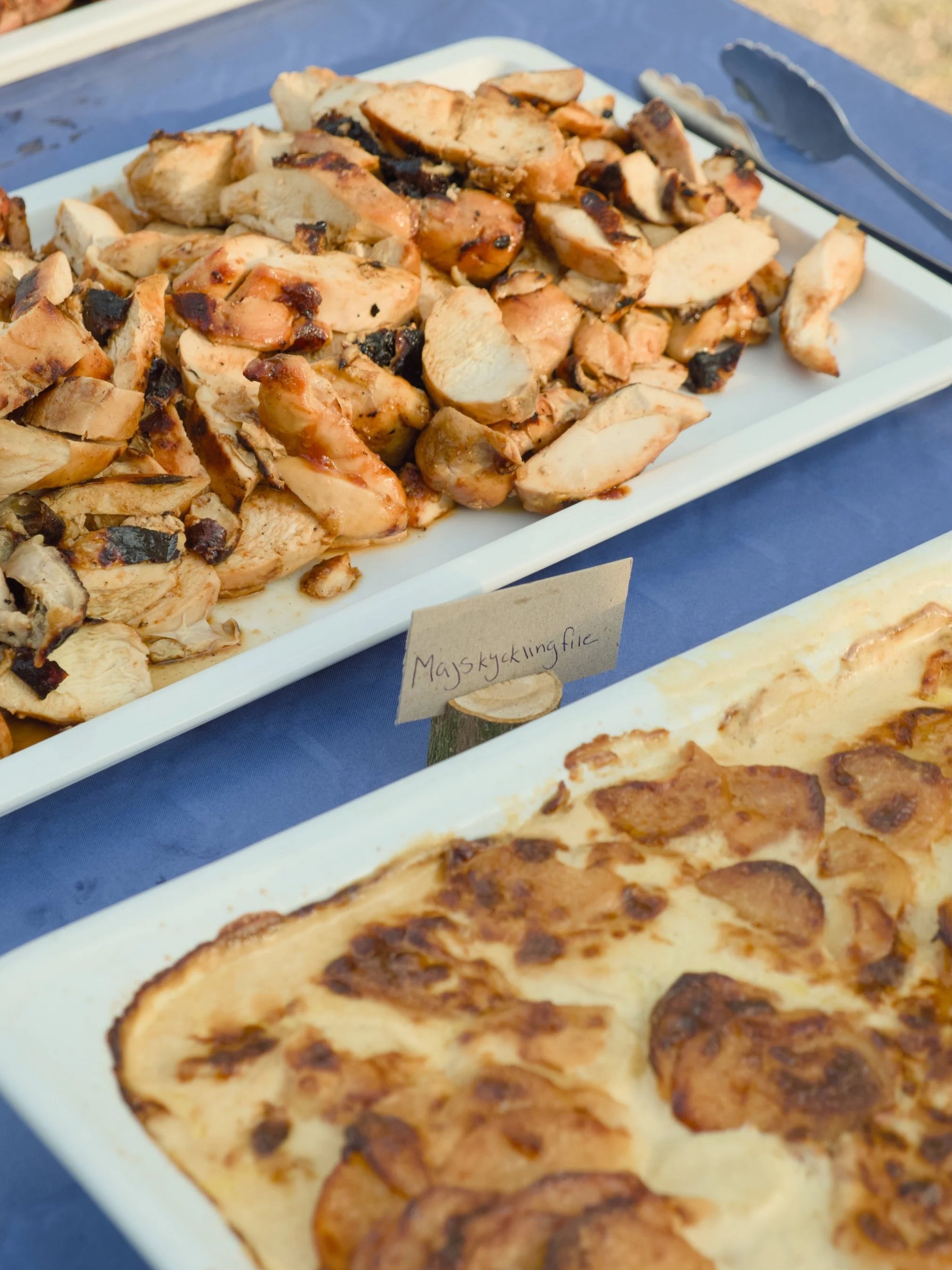Cooked chicken pieces on a white platter with a small handwritten sign in Swedish beneath it, sitting on a blue tablecloth. Part of a baked dish, possibly a potato gratin, is visible in the foreground.