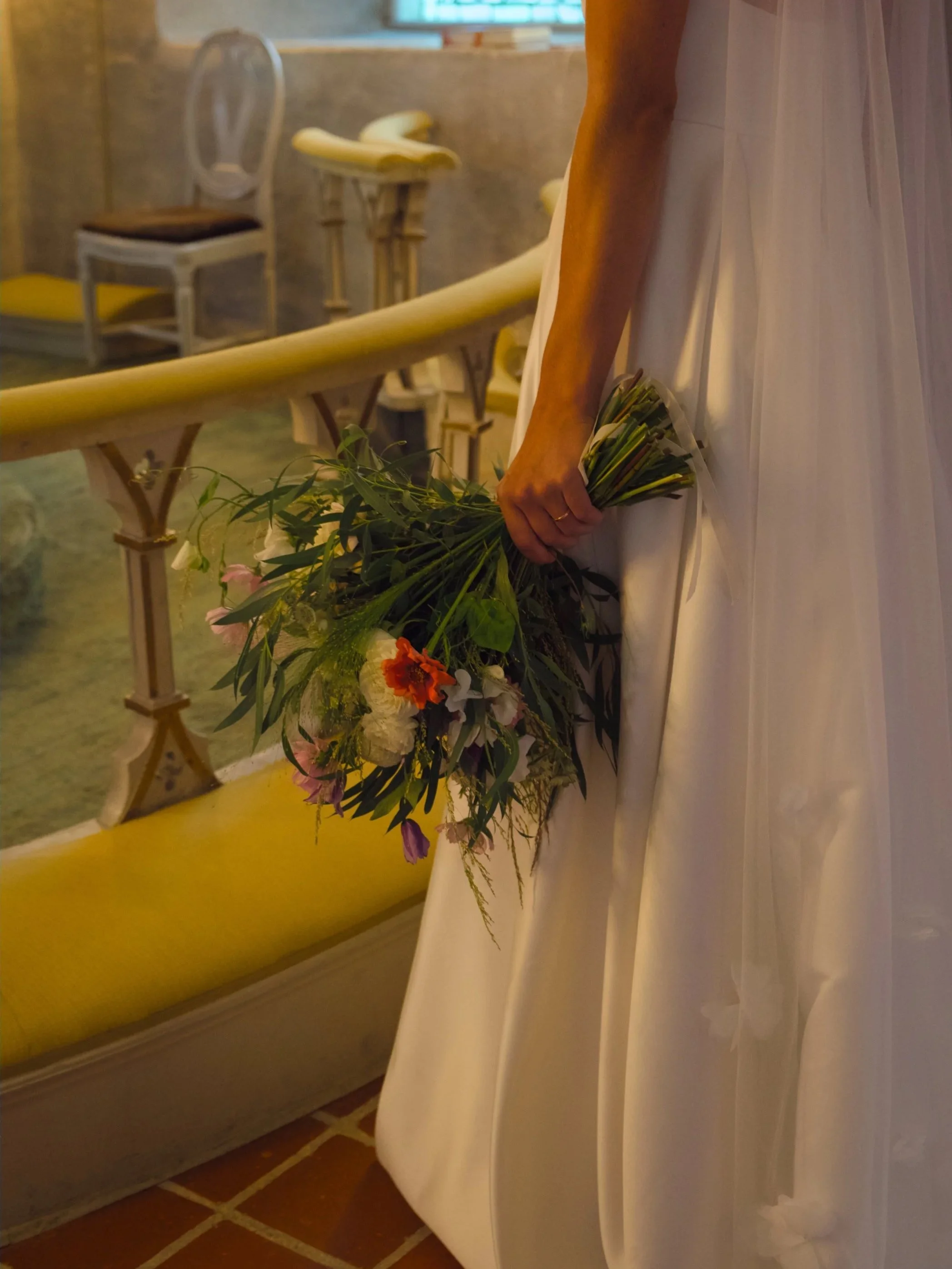 A bride holding a bouquet of flowers in a white wedding dress, standing in a softly lit, rustic indoor setting.