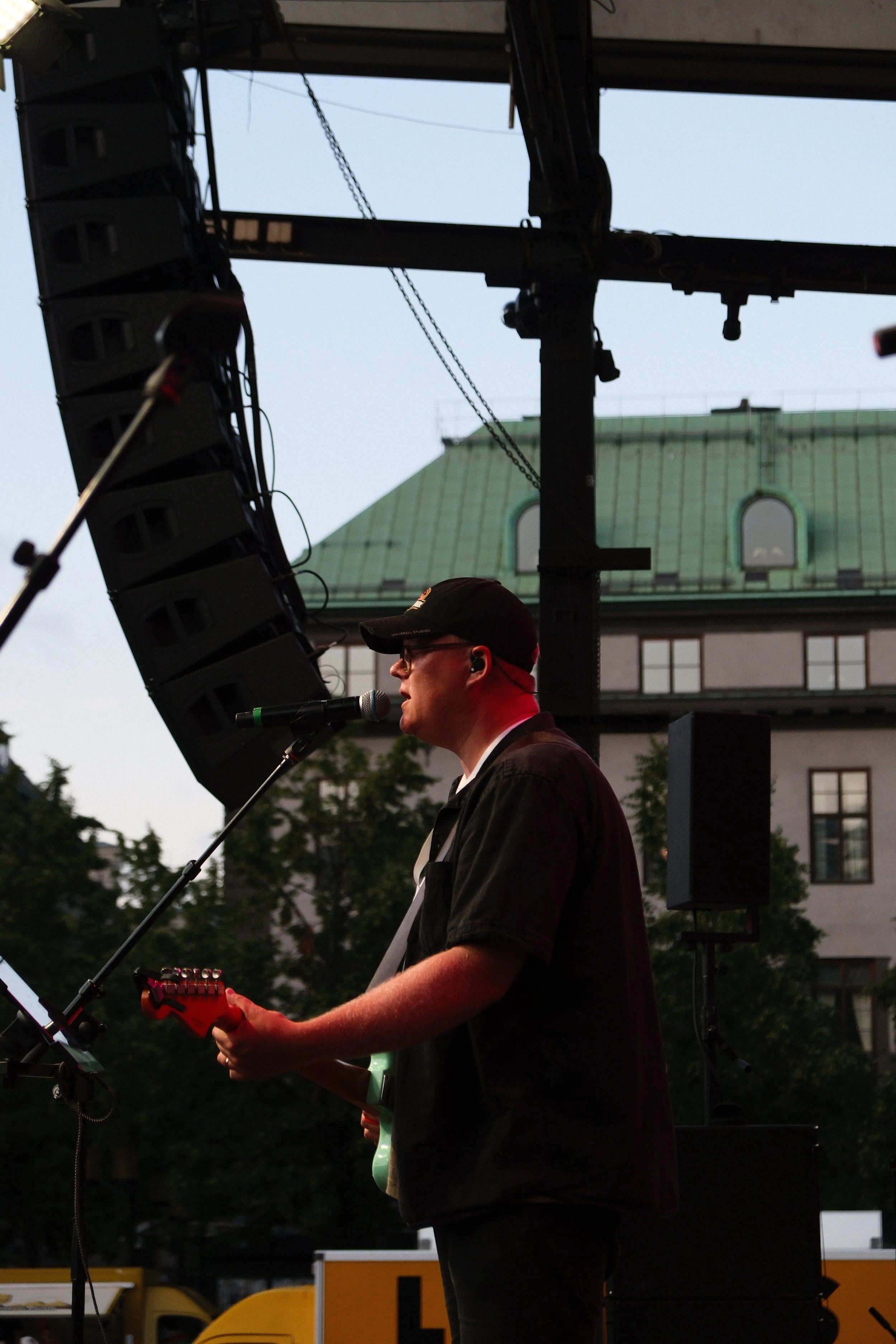 A musician playing an electric guitar and singing into a microphone on stage outdoors during evening.