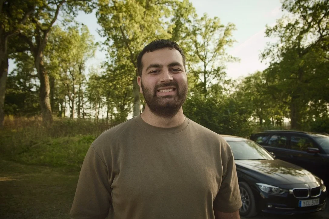A smiling man with a beard wearing a brown T-shirt standing outdoors with trees and cars in the background during daytime.