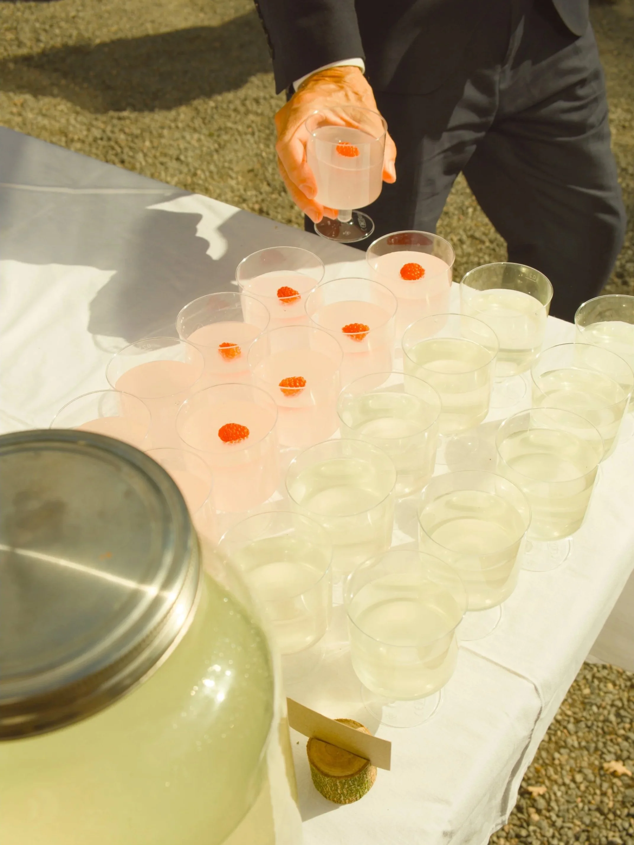 A man in a suit pouring a clear beverage with red berries in clear wine glasses on a table covered with white cloth.
