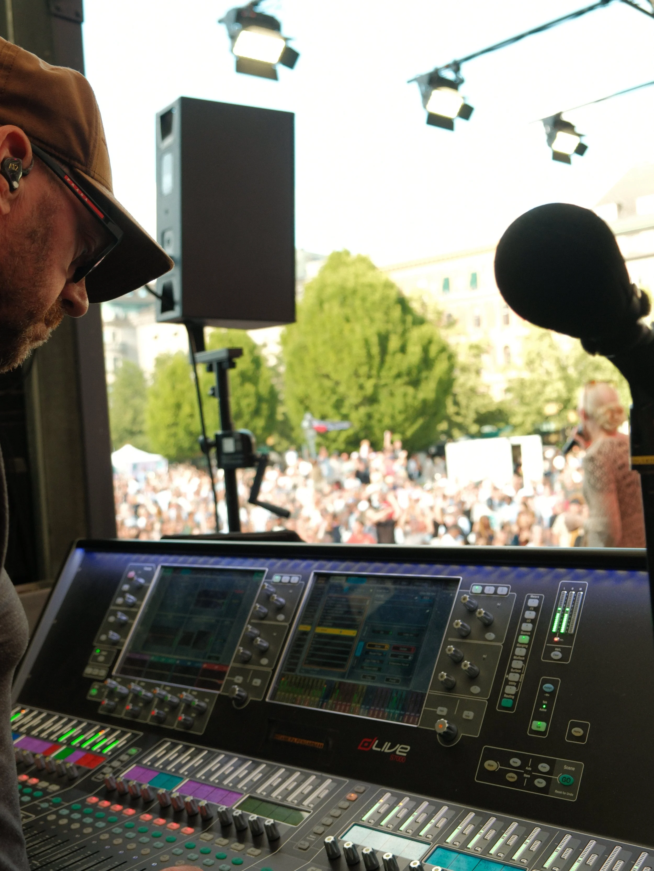 A man operating a digital sound mixing console outdoors during a live event, with a large crowd and green trees in the background.