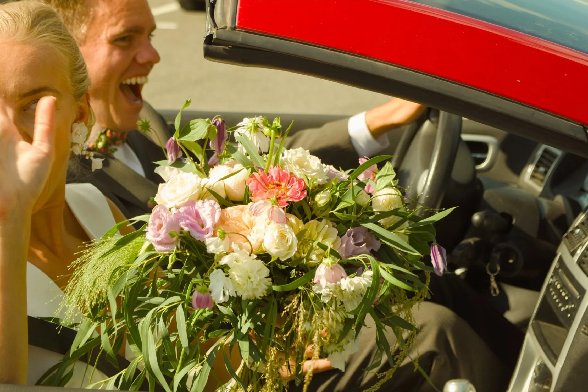 A bride and groom in a car, holding a large bouquet of flowers, celebrating their wedding.