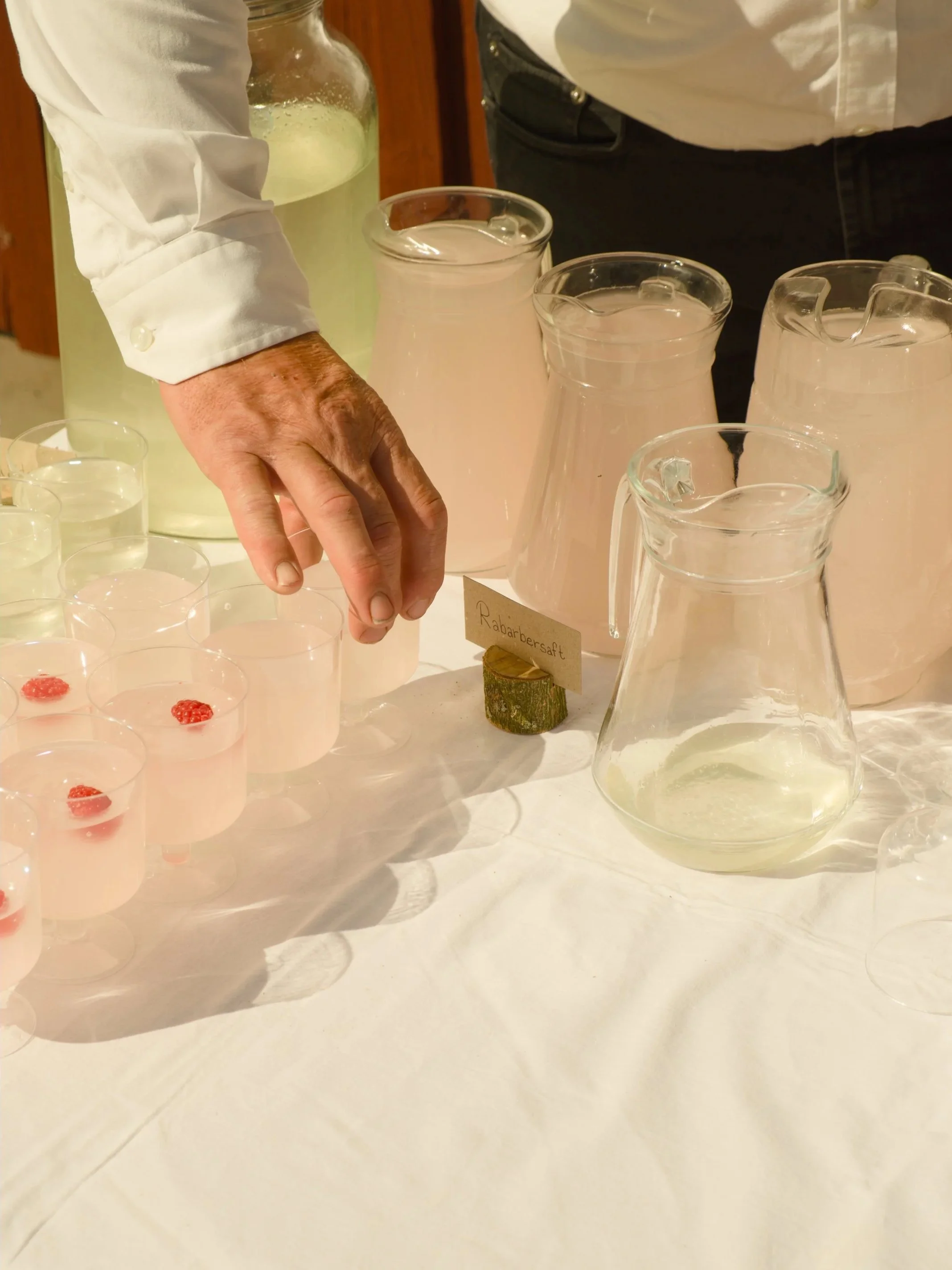 A person in a white shirt standing behind a table with several glass pitchers and cups filled with a pink liquid, some garnished with raspberries, and a small sign with the word 'Rhabarbersaft', indicating this is a setup for serving rhubarb juice.