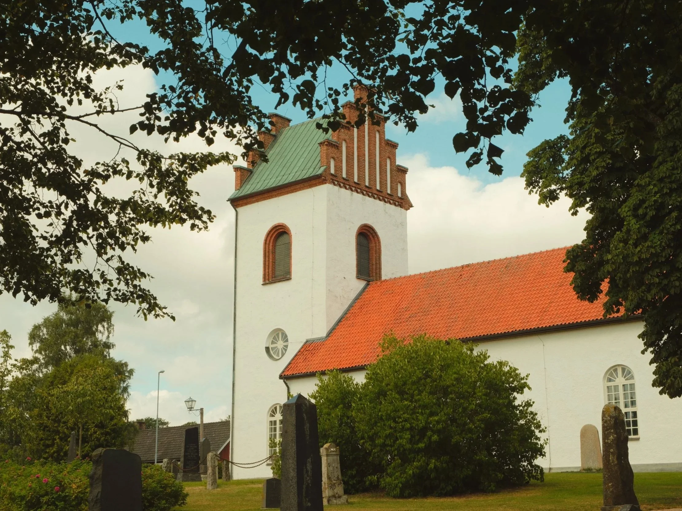 A white church with a green roof, red-tiled roof section, and a tall steeple, surrounded by trees and grave markers in the churchyard.