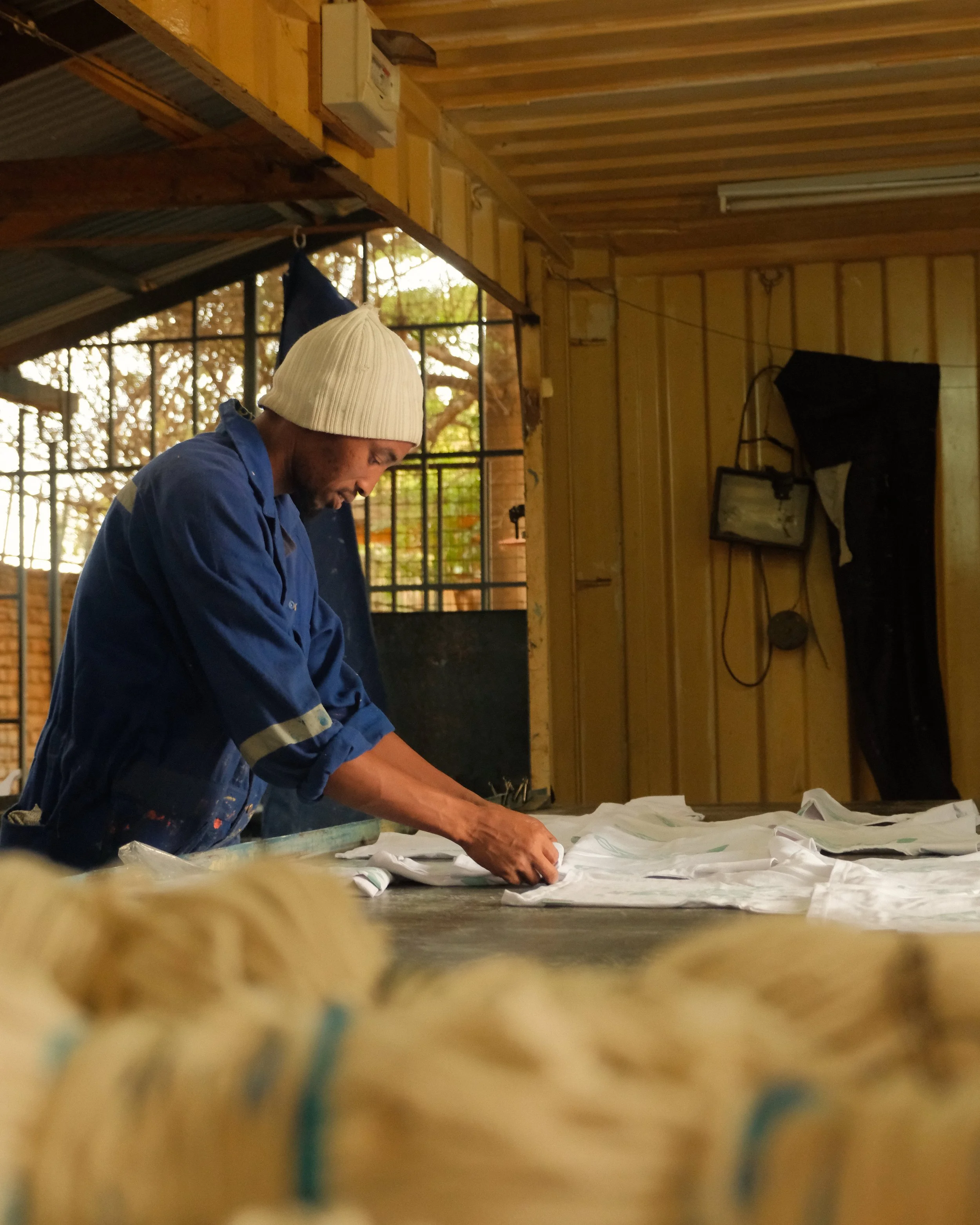 A man wearing a white beanie and blue work shirt is working with white fabric or clothing on a workbench inside a wooden workshop.