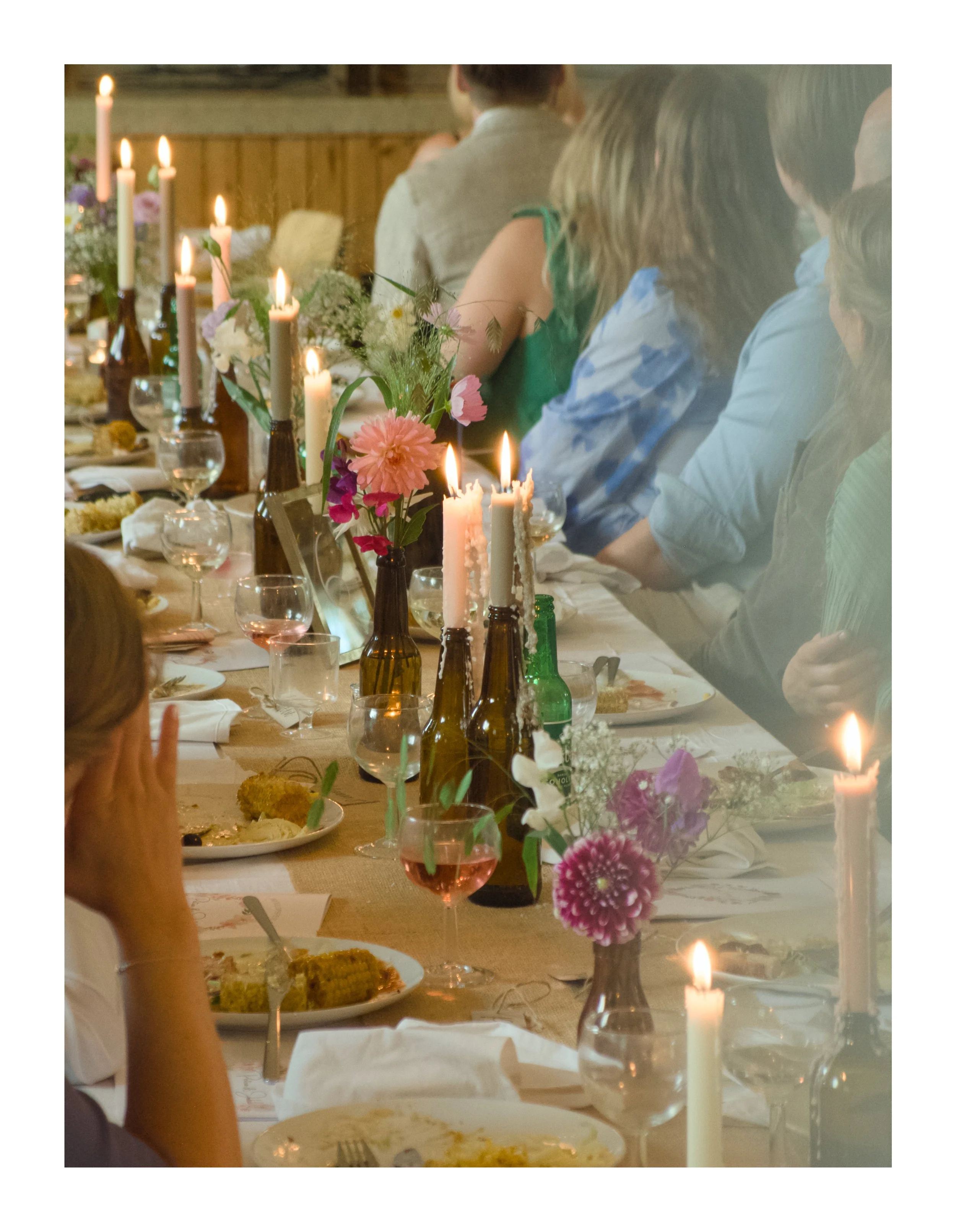 A family or group of people seated at a decorated dining table with lit candles, flowers in vases, and glasses of pink drink, likely celebrating a special occasion.