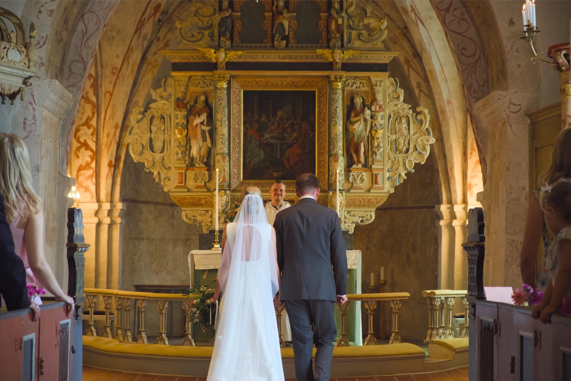 A bride and groom standing at the altar during a wedding ceremony in a church, facing a priest or officiant, with guests observing on the sides.