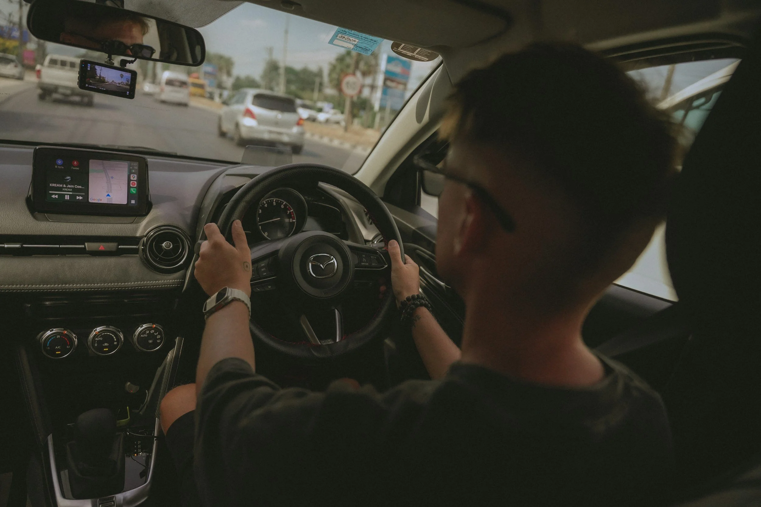 A person driving a Mazda car on a busy road, with traffic and billboards visible through the windshield.