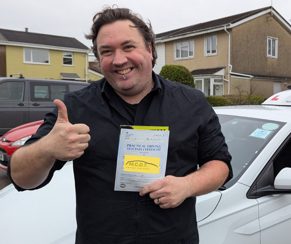 A man is standing outdoors next to a white car, holding a practical driving test pass certificate, and giving a thumbs-up while smiling.
