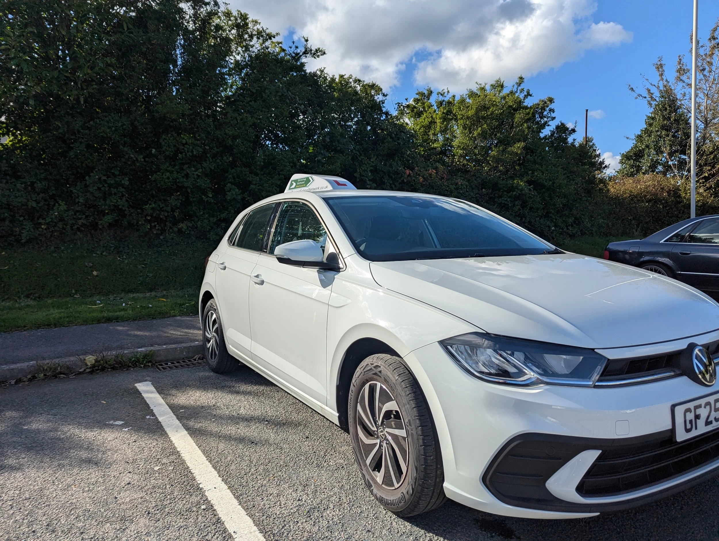 White Volkswagen car parked in a parking lot on a sunny day with trees and a partly cloudy sky in the background.