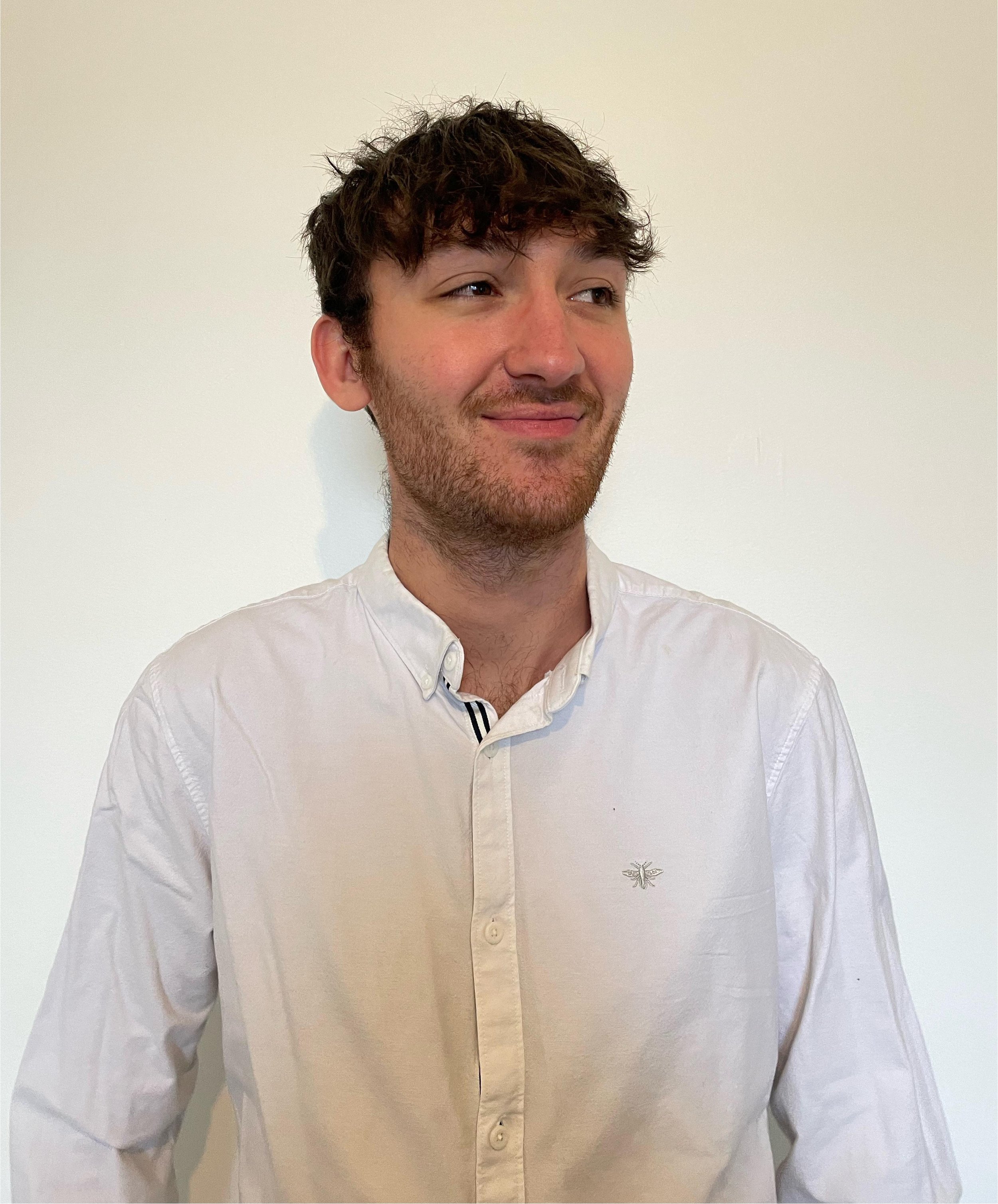 A young man with curly brown hair, a beard, and mustache, wearing a white button-up shirt with an embroidered bee logo, standing against a plain white wall, looking to the side with a slight smile.