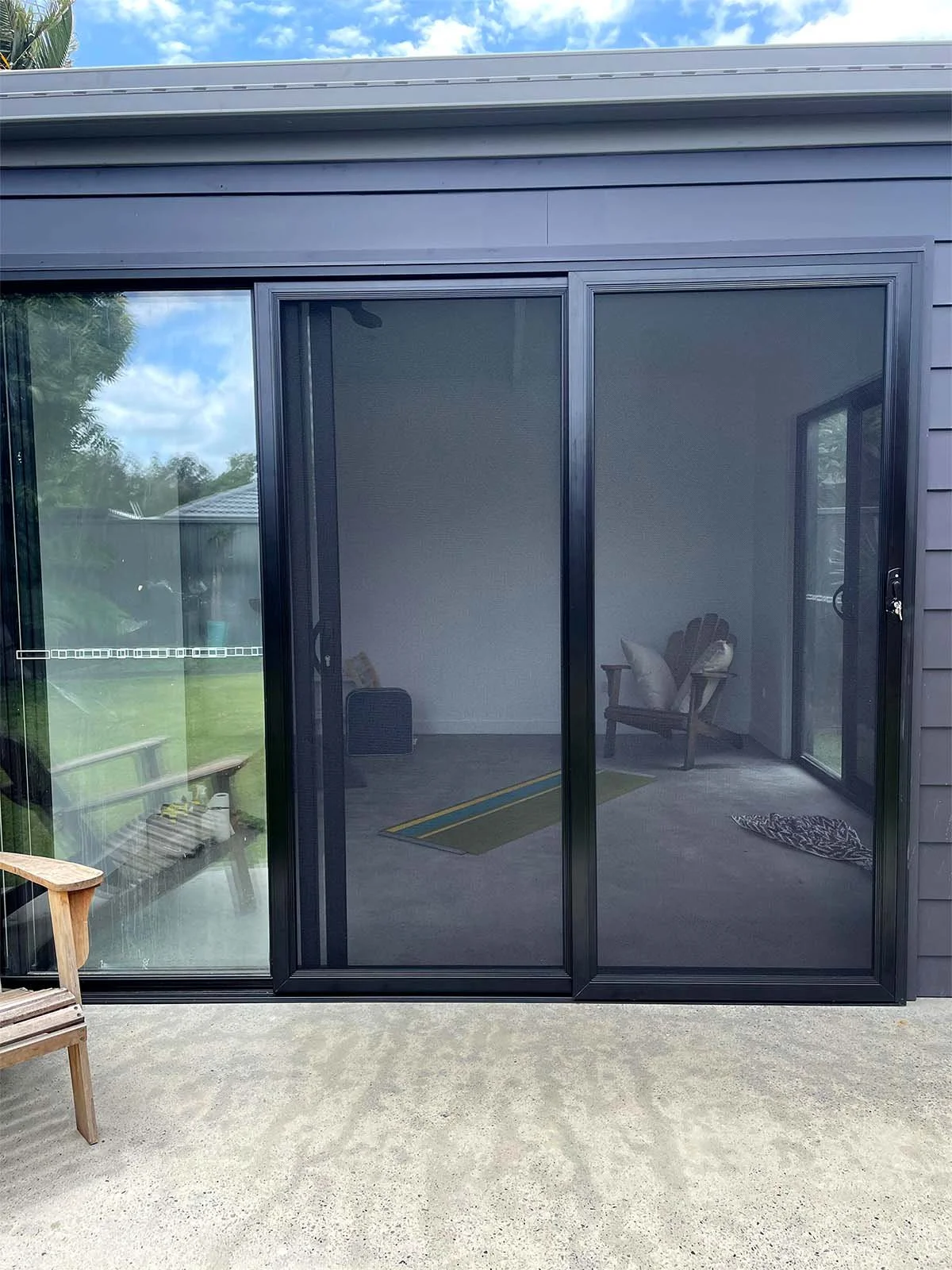 Security screen sliding door installation Southeast Queensland on a house with an outdoor patio, with a view of the interior room showing a wooden chair with pillows, a rug, and a visible Window.