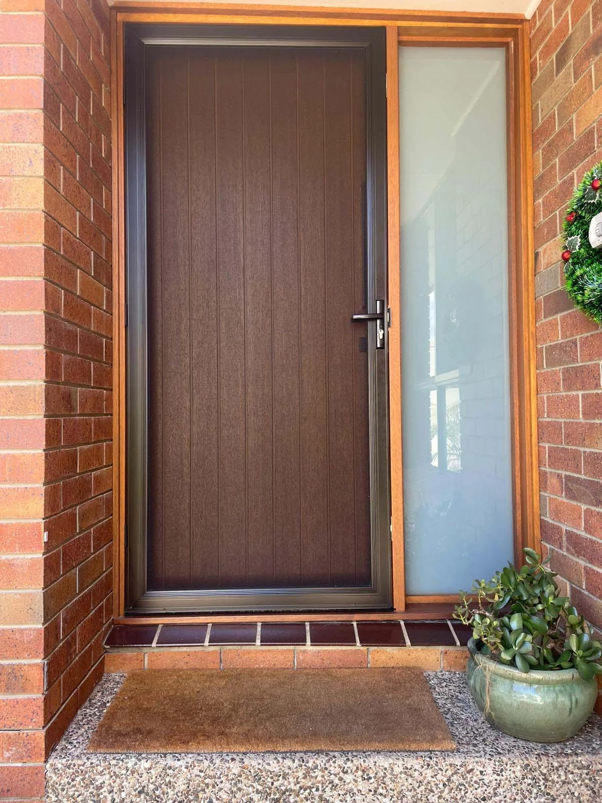 Screenguard accredited installation of balck security screen front door with a brown textured door and a glass side panel, brick walls, a brown doormat, and a potted plant nearby.