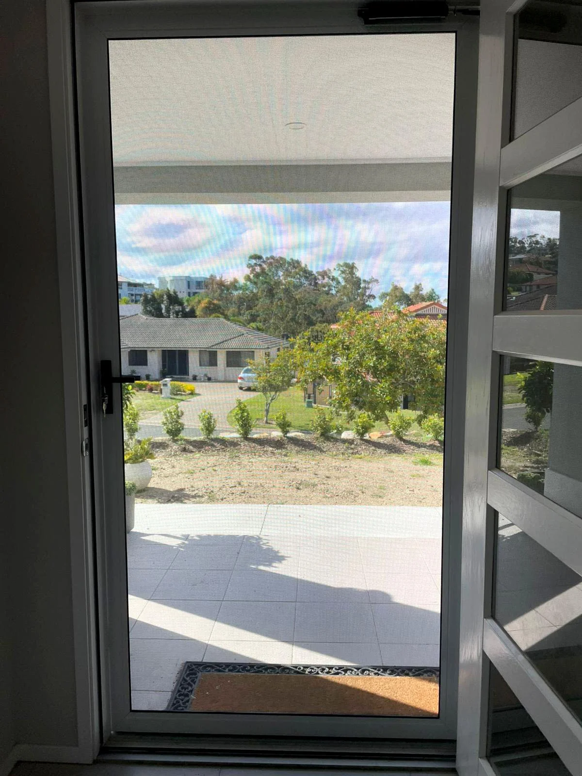 View through a security screen door onto a patio with tiled flooring, a garden with shrubs and trees, houses in the background, and a partly cloudy sky.