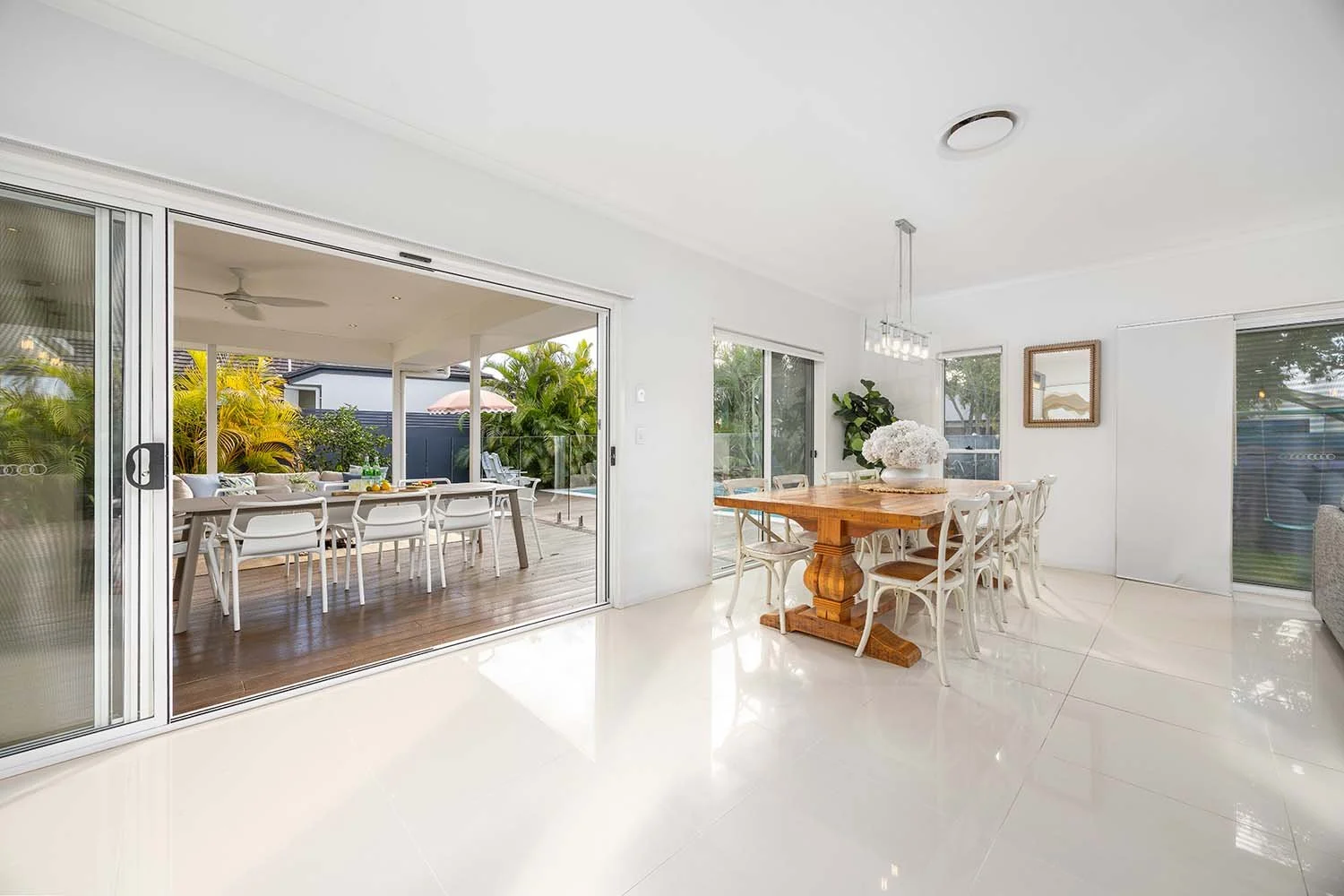 Modern dining room with white walls and tiled floor, featuring a wooden dining table and a white security sliding door leading to outdoor patio with additional seating, and greenery outside.