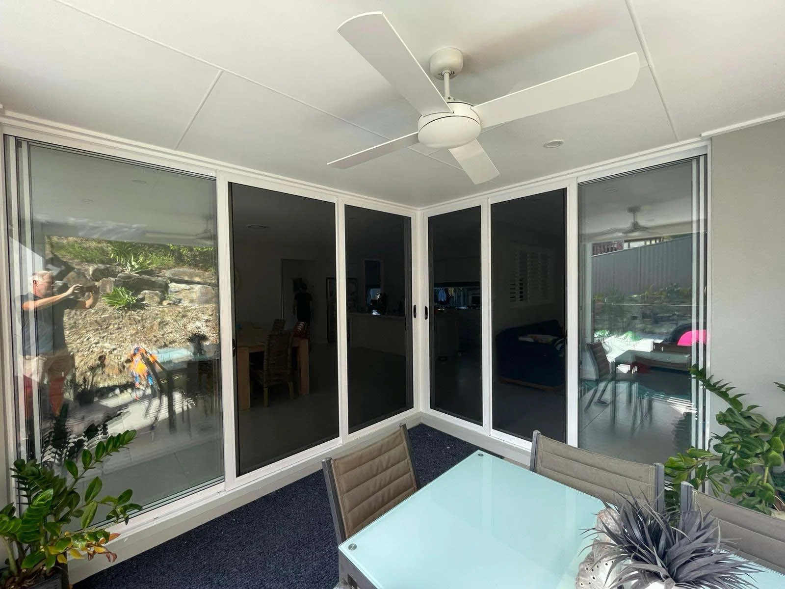 Outdoor patio with white security screen doors, ceiling fan, glass table, brown chairs, and potted plants. Located at Tugun on the Gold Coast.