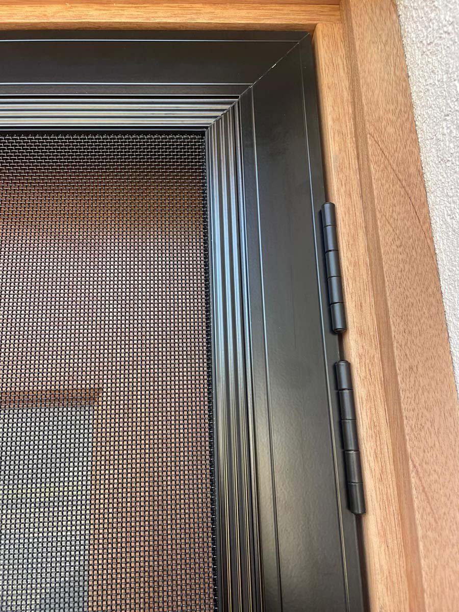 Close-up of a window with a Stainless steel security screen door black mesh screen, black hinges, and a wooden frame on the right side.
