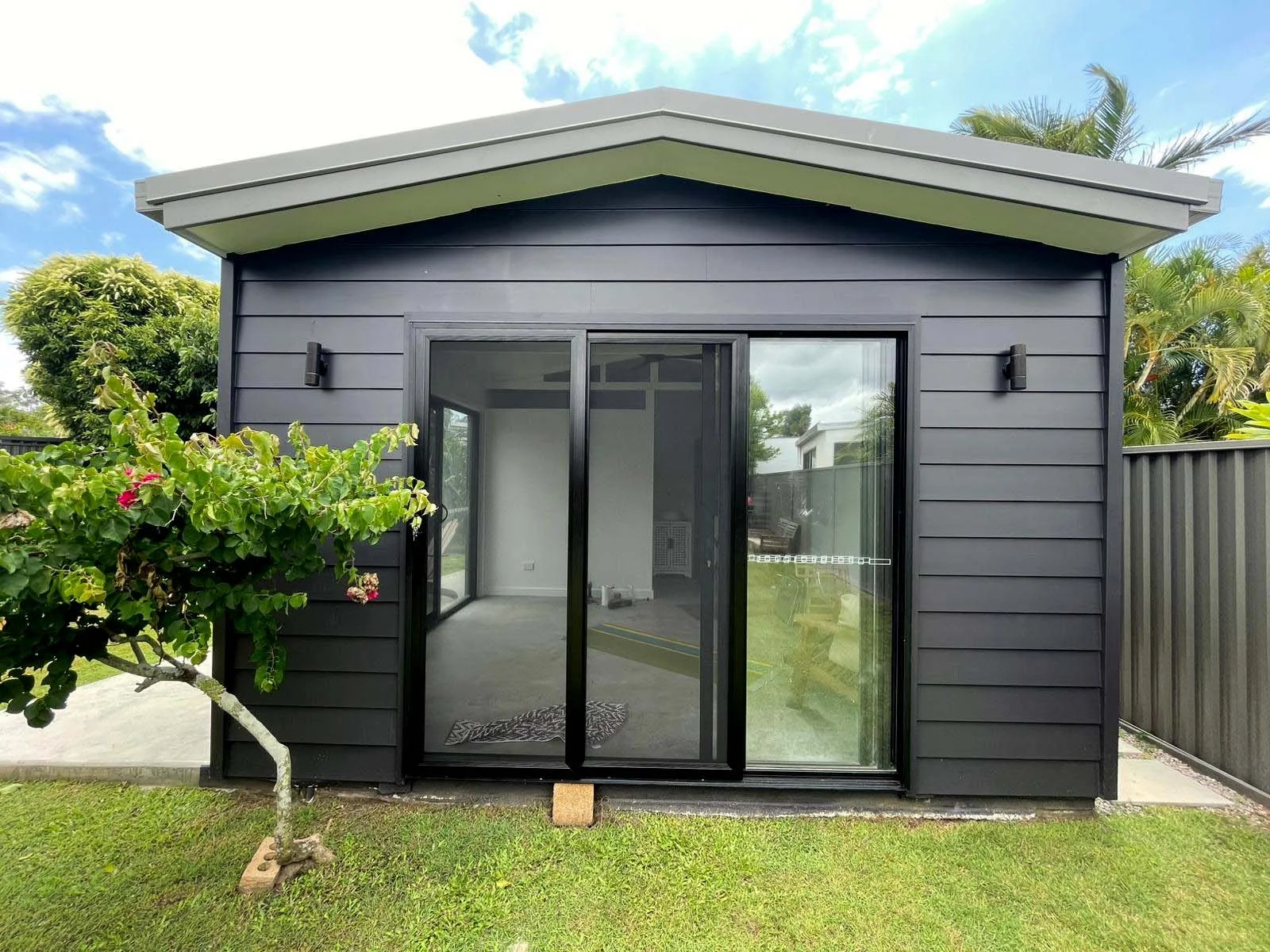 Black stainless steel security door with triple lock Gold Coast on a granny flat surrounded by green grass, trees, and a wooden fence, under a partly cloudy sky.
