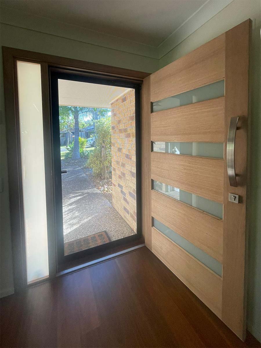 View of a front door with a modern wooden slatted design and glass panels, opening to a security screen outside with greenery and a brick wall.