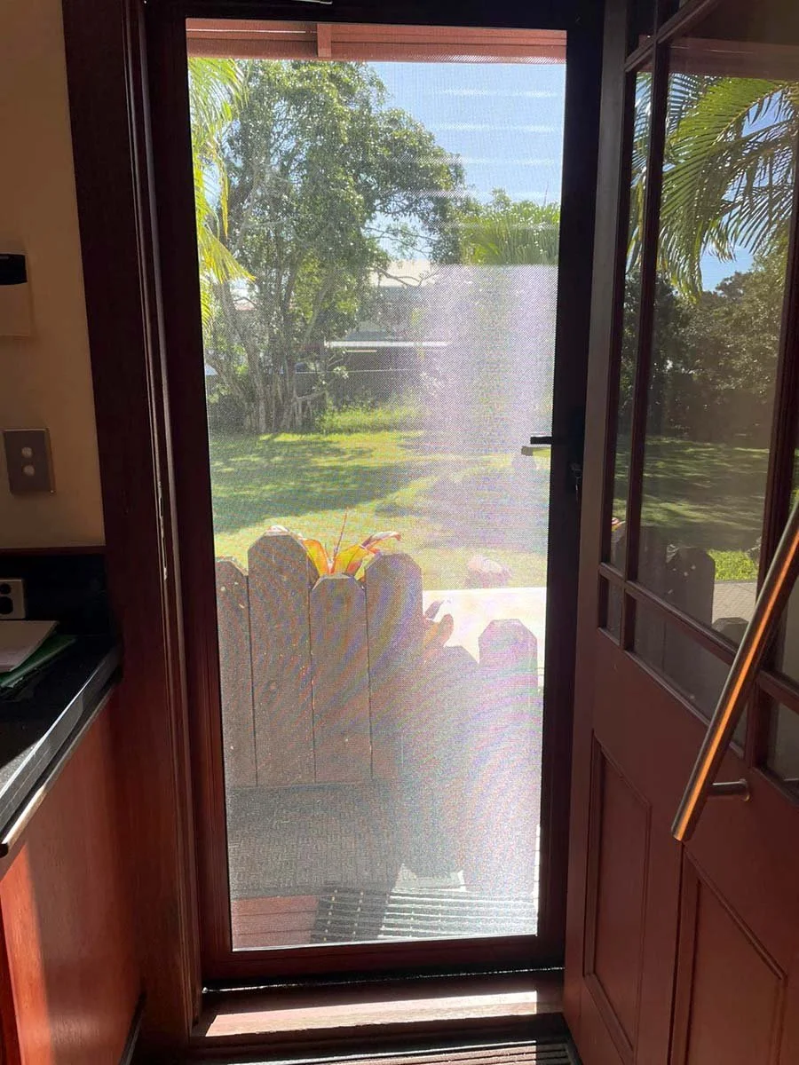 View through a security door showing a wooden railing with potted plants, green trees, and a sunny yard.