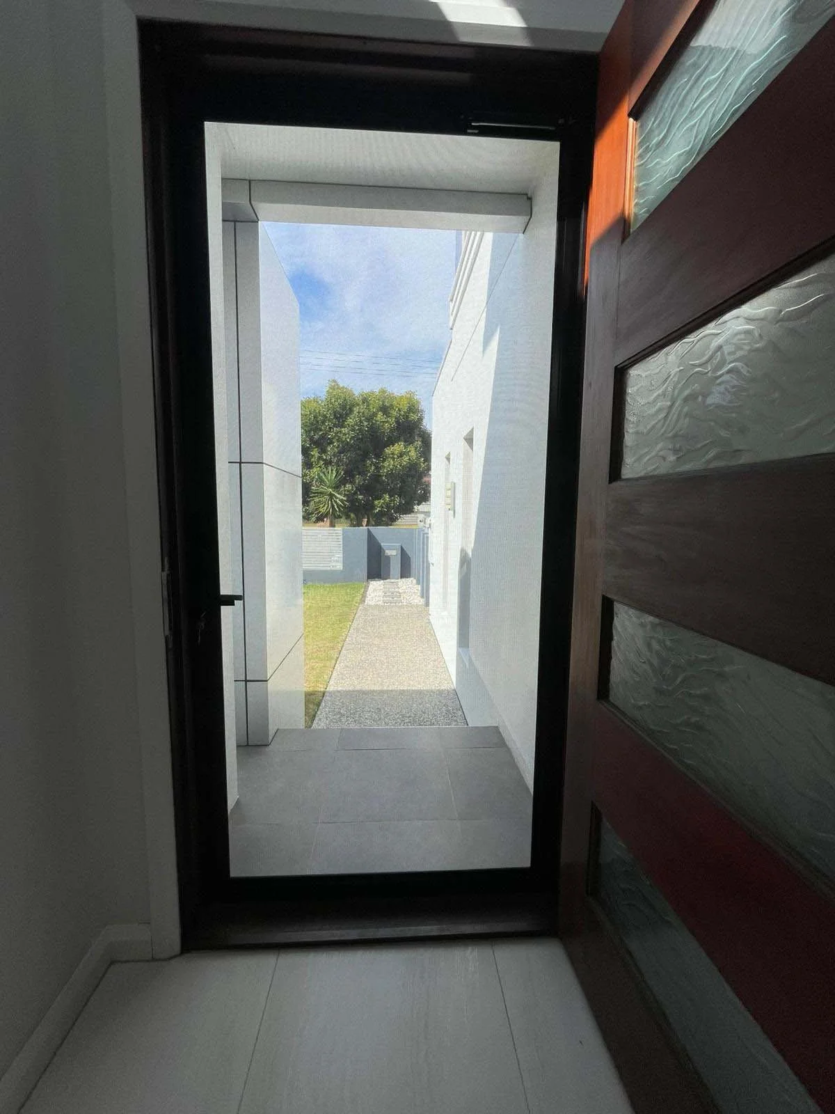 View through a Stainless steel security screen door installation Gold Coast home showing an outdoor pathway with grass, white buildings, and a tree against a blue sky.