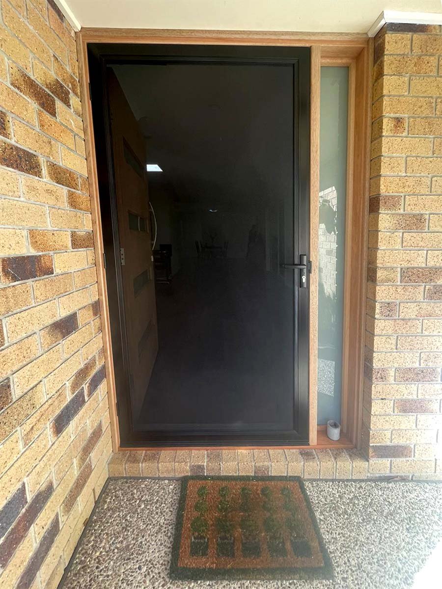 View of a black screened front door with a wooden frame, flanked by a small glass side panel, opening to a brick porch with a doormat featuring a green and black triangle design. Security screen door installation Gold Coast