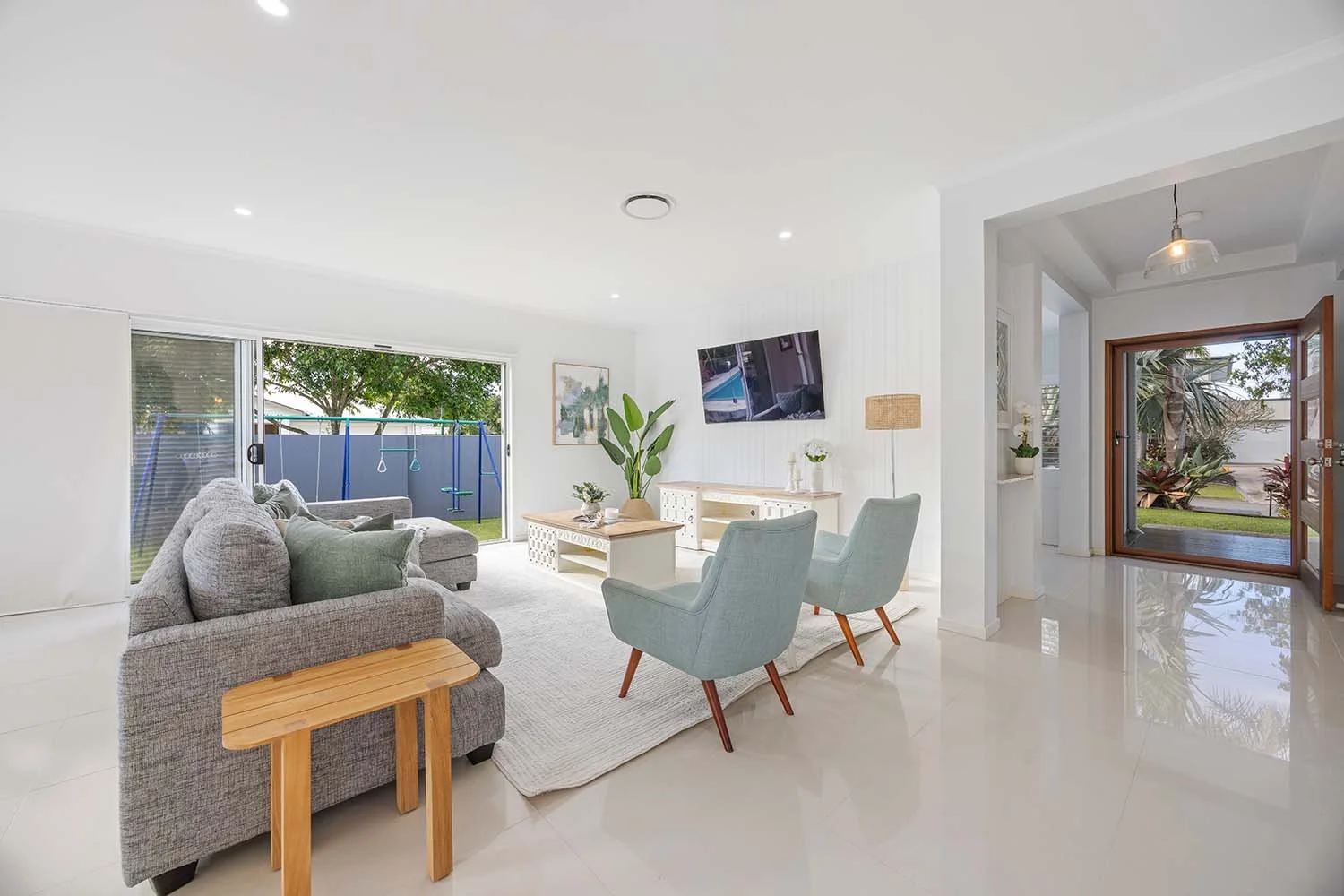 Bright living room with large sliding security door leading outside to backyard with swings and greenery. Inside there is grey sofa , two green armchairs,  and TV in living room.
