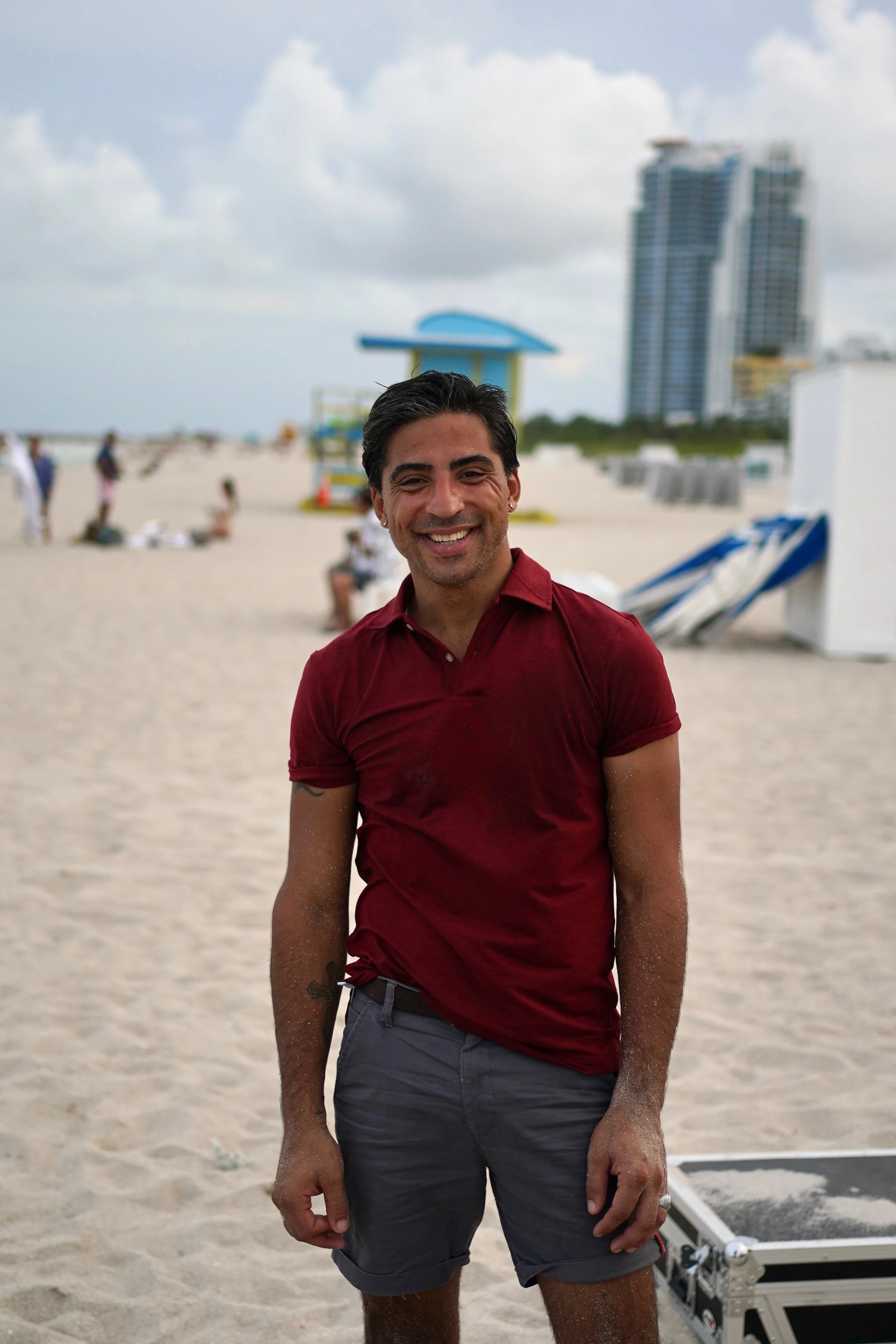 A smiling man in a red shirt and gray shorts standing on a sandy beach. There are blurred people, a lifeguard tower, and high-rise buildings in the background under cloudy skies.