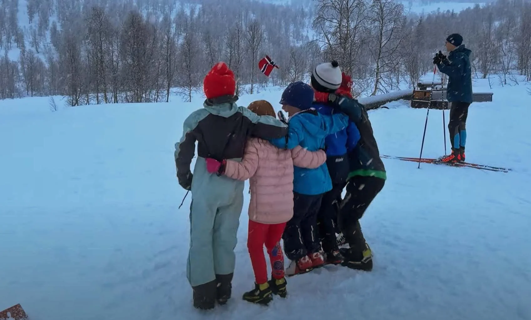 Gruppemedlemmer i snøen som holder rundt hverandre og heier, mens en person på ski tar bilder i bakgrunnen.