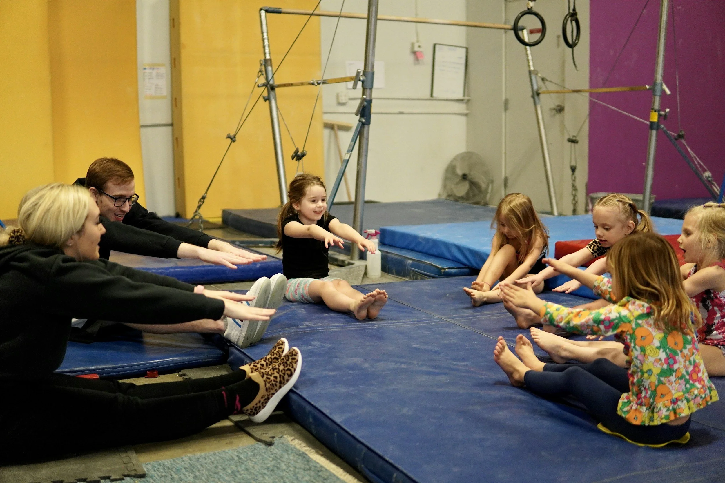 Barn og voksne sitter på yogamatte i sirkelform, øver på å strekke armene framover med bena utstrakt i en gymnastikkøvelse. Bakgrunn med turnutstyr og gulvegg.