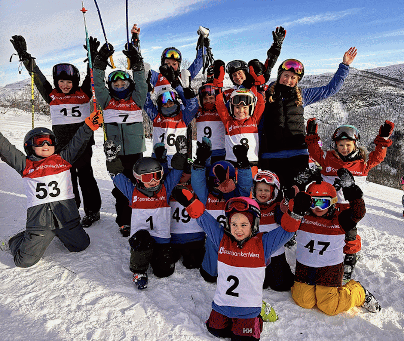 En gruppe barn og voksne i skihjelmer og skiklær på snø, feirer og smiler mot en snødekket fjellbakgrunn.