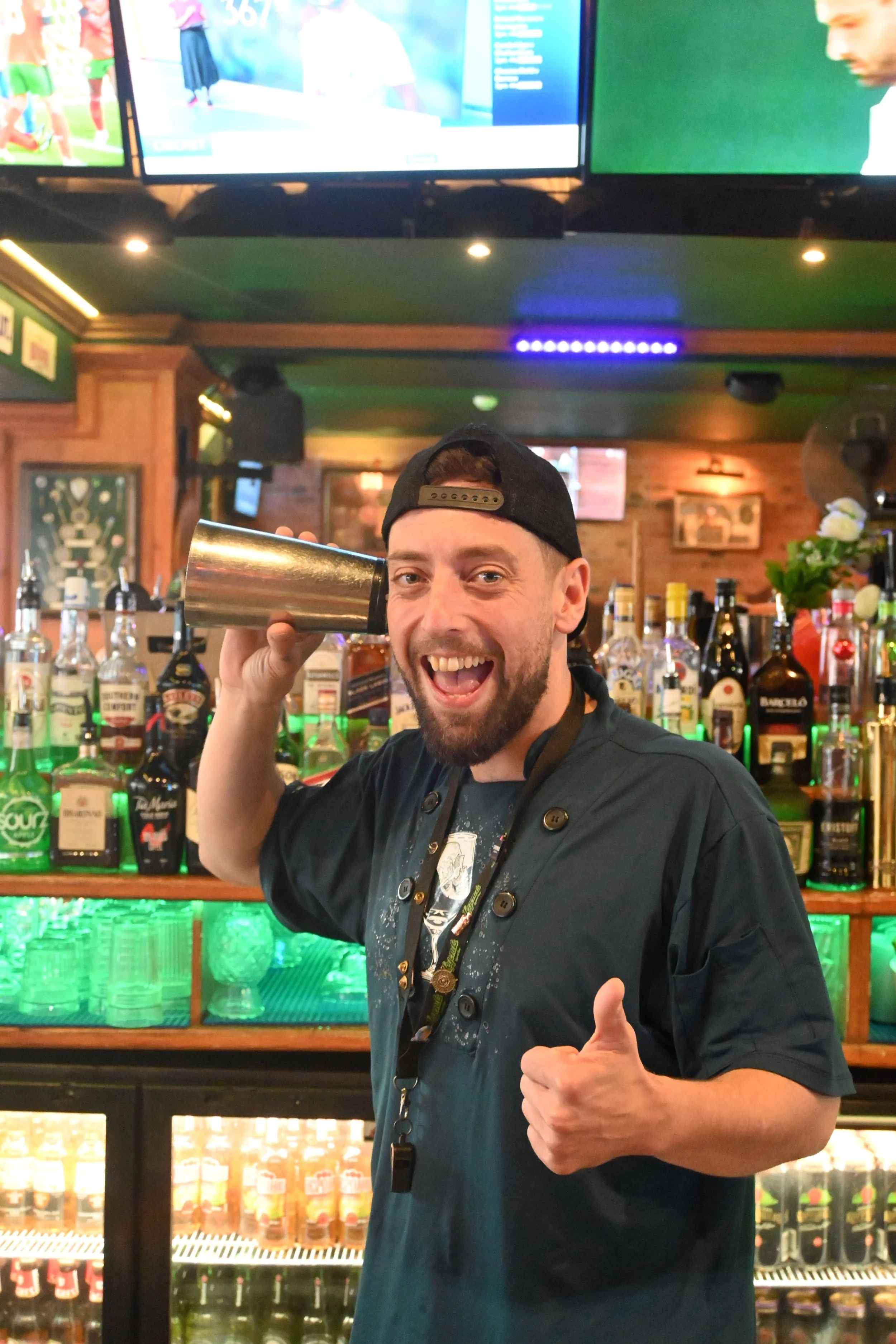 A bartender with a beard, wearing a black cap backward and a black shirt, is smiling and holding a metal cocktail shaker to his ear. He is giving a thumbs-up with his other hand in a bar with various bottles of alcohol behind him and a television screen above.