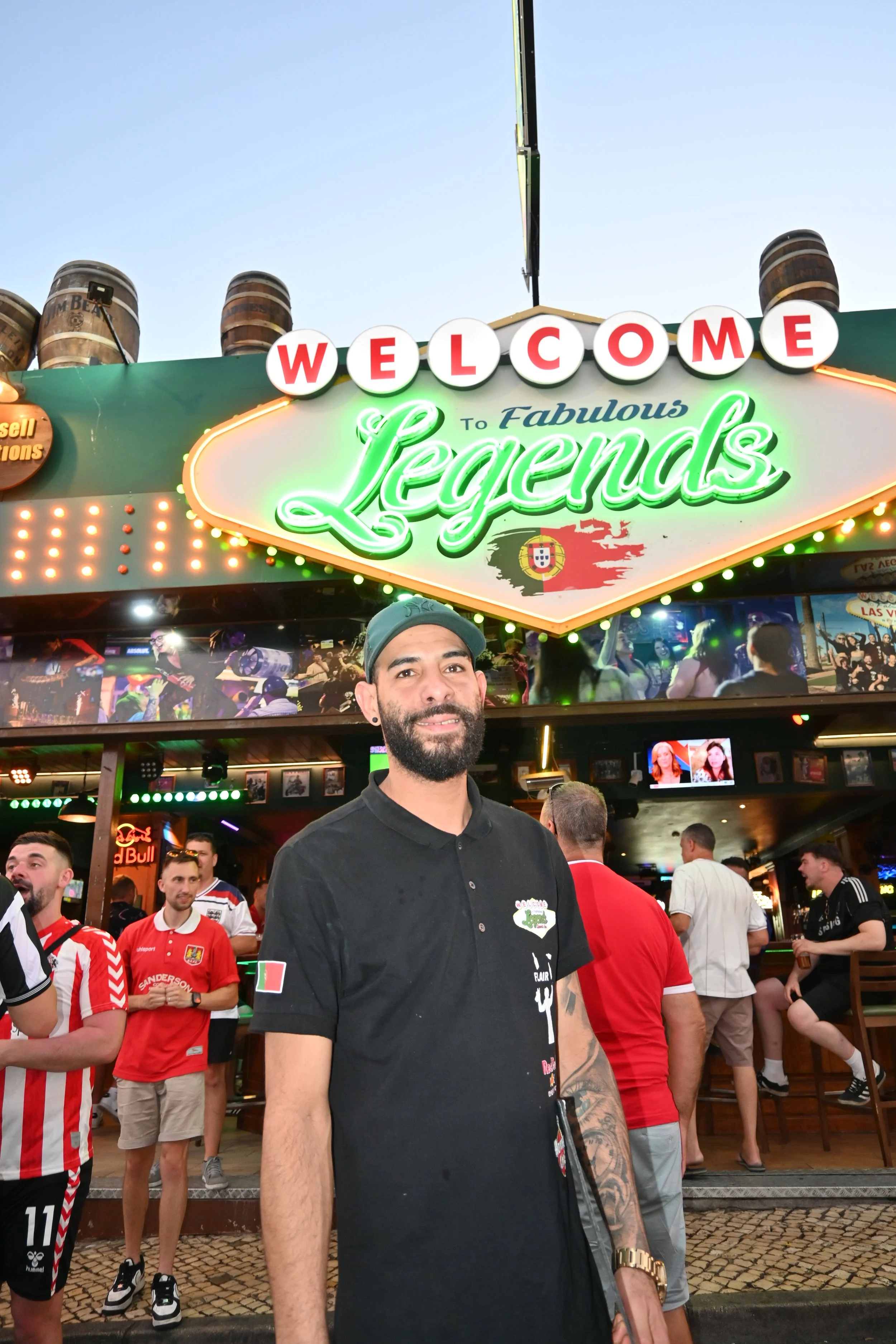 A man standing at a sports bar or stadium event with a large illuminated sign reading 'Welcome to Fabulous Legends' and several people wearing sports jerseys around him.