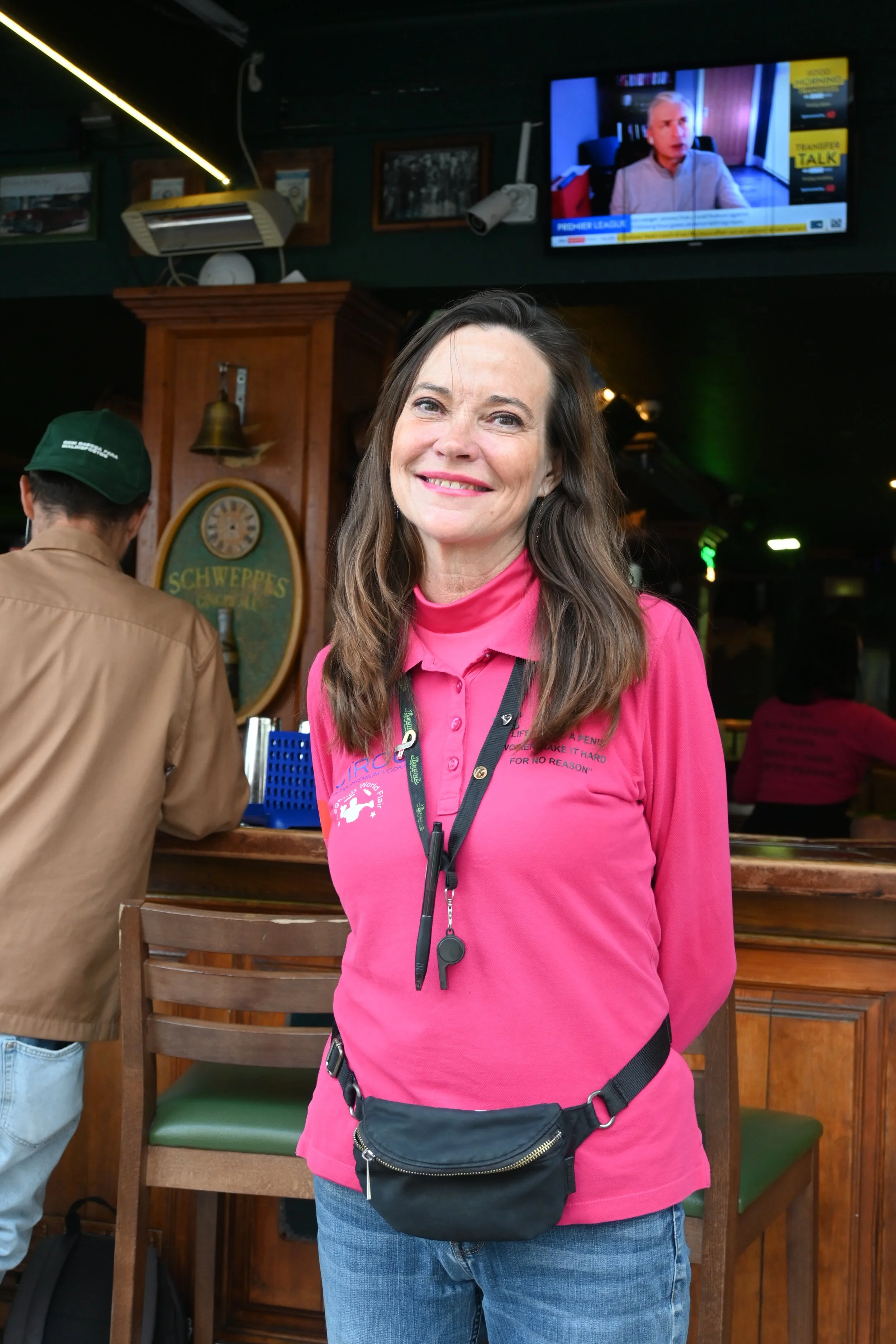 Smiling woman with long brown hair wearing a pink long sleeve shirt and a black waist bag standing in a restaurant or bar with wooden furniture and a television screen in the background.