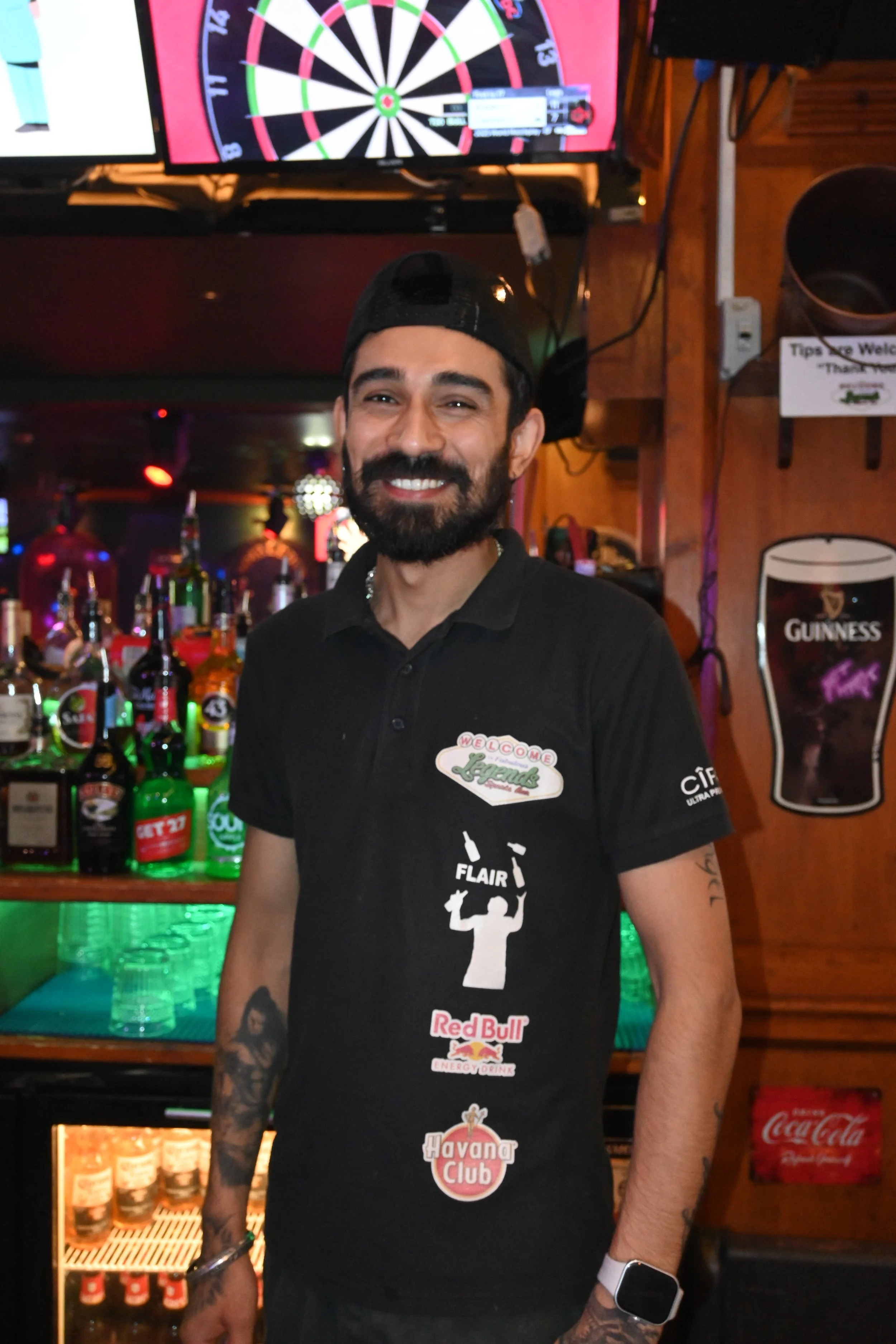 A man with a beard and tattoos standing in front of a bar with various bottles of alcohol, wearing a black t-shirt with sponsor logos, smiling. Behind him, there is a dartboard on the wall and a Guinness beer poster.