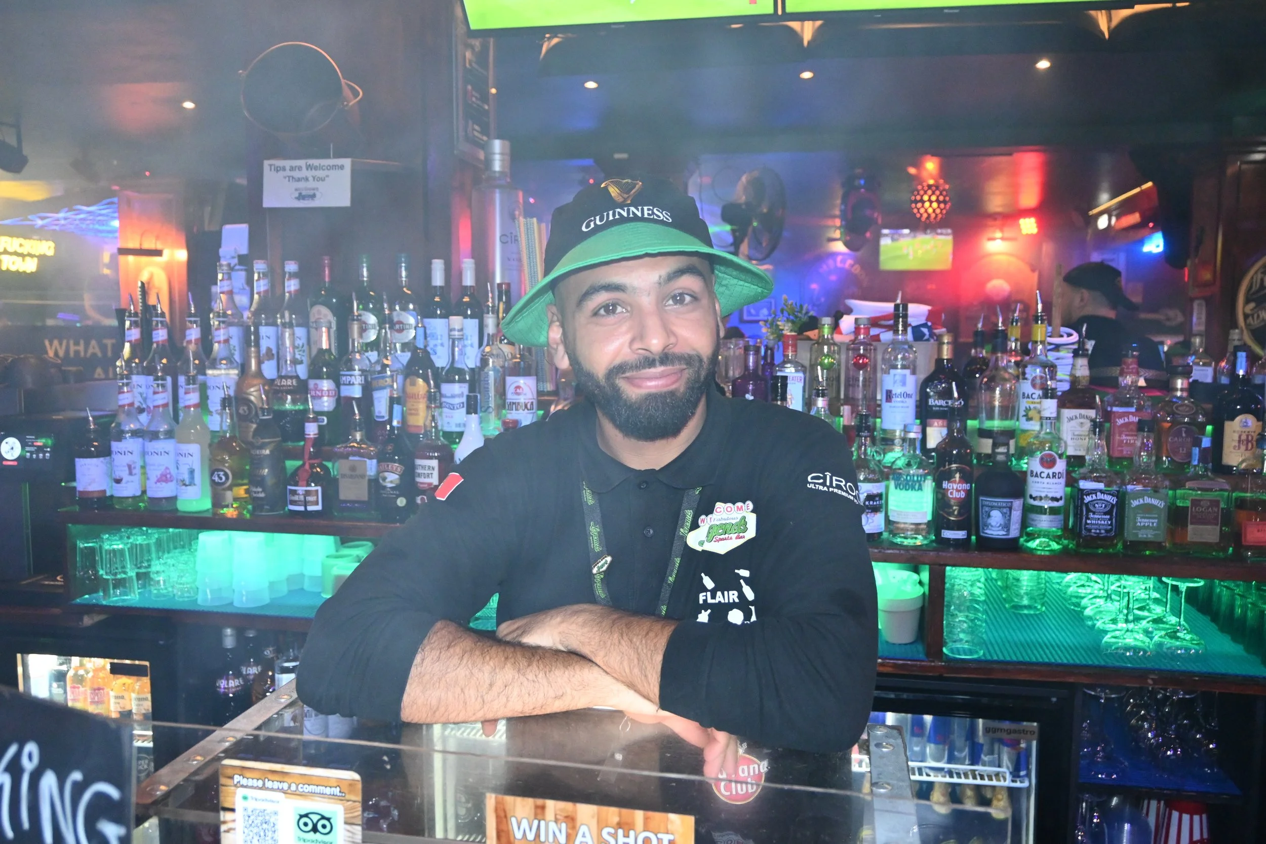 A bartender with a beard and a Guinness hat standing behind a bar with assorted liquor bottles, neon lighting, and a TV screen in the background, in a lively bar setting.