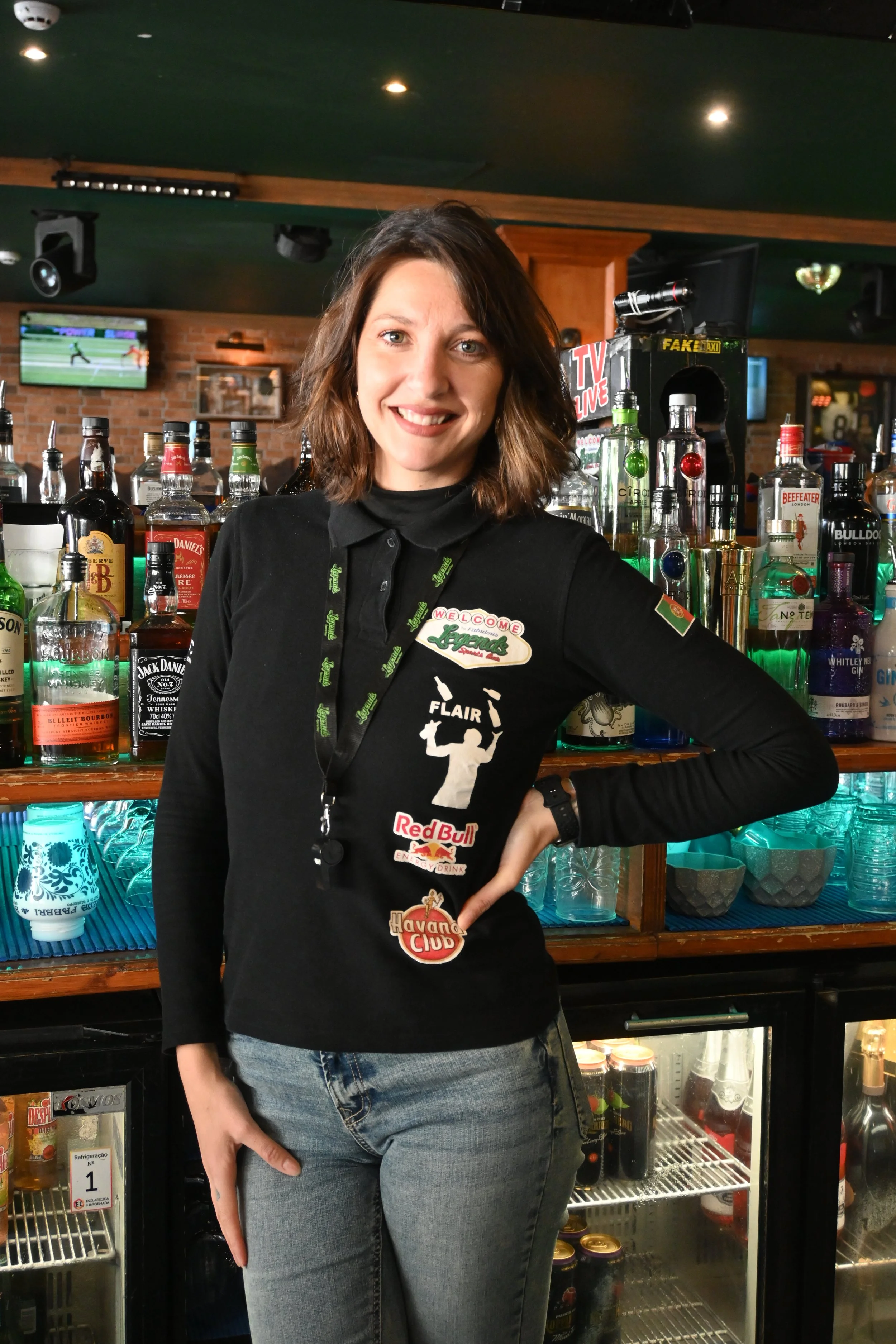A smiling woman standing behind a bar with various liquor bottles and glasses, wearing a black shirt with logos, in a dimly lit pub or bar setting.