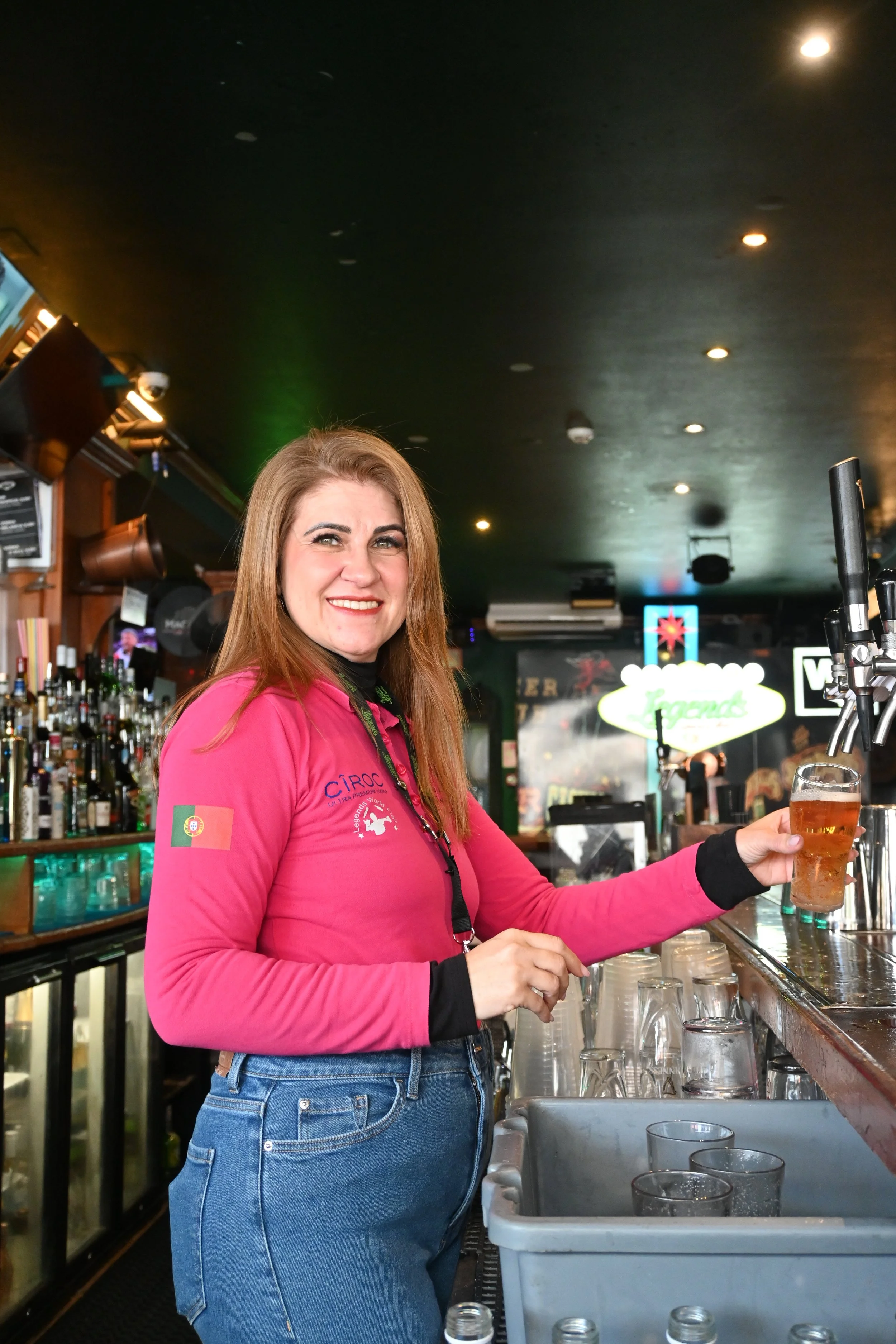 A woman with long red hair and a pink jacket standing at a bar, pouring a drink into a glass.
