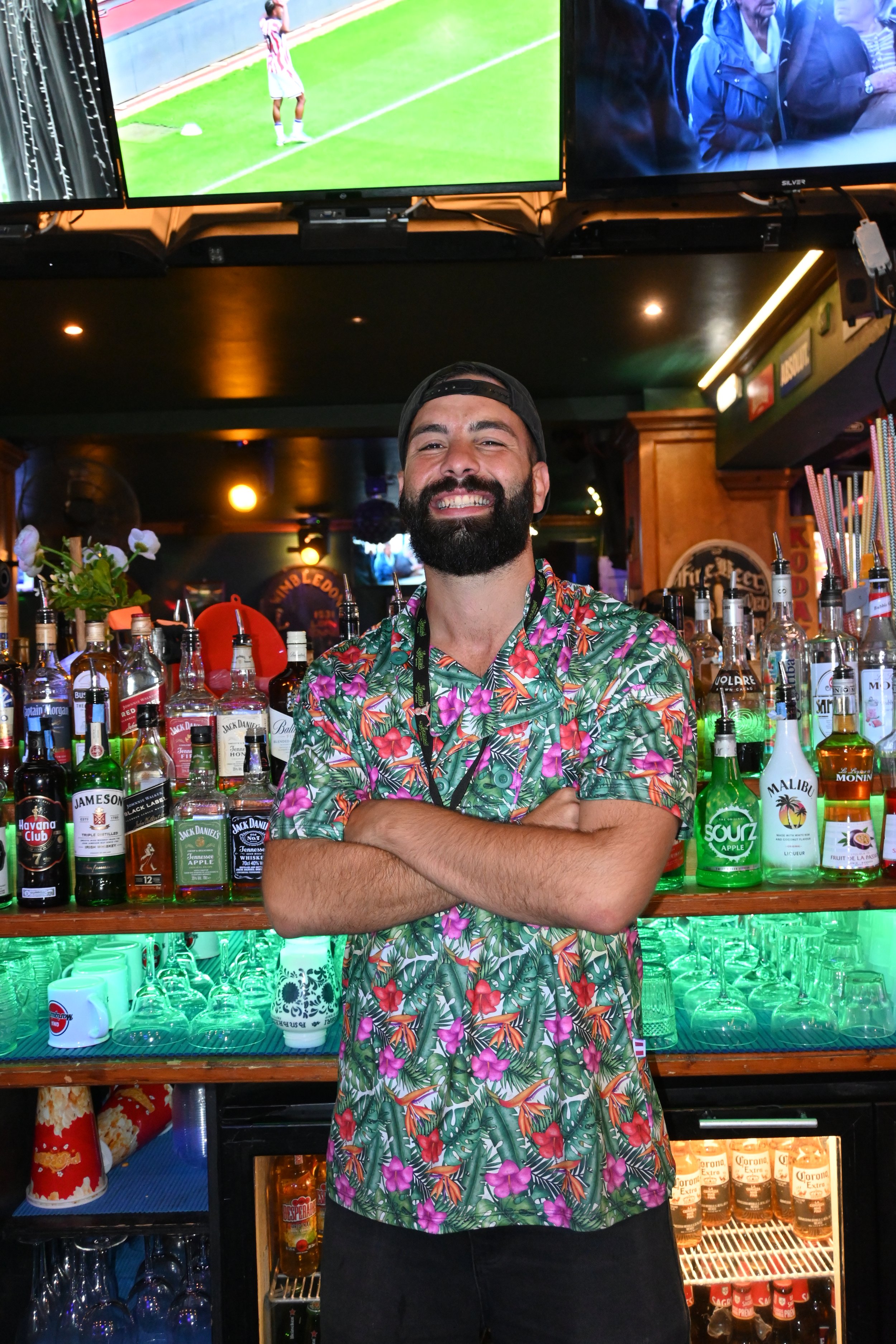 A smiling man with a beard and a black cap standing behind a bar, wearing a colorful hawaiian shirt. The bar is stocked with various bottles of liquor, and there are multiple TVs mounted above showing different sports games and scenes in a lively bar setting.