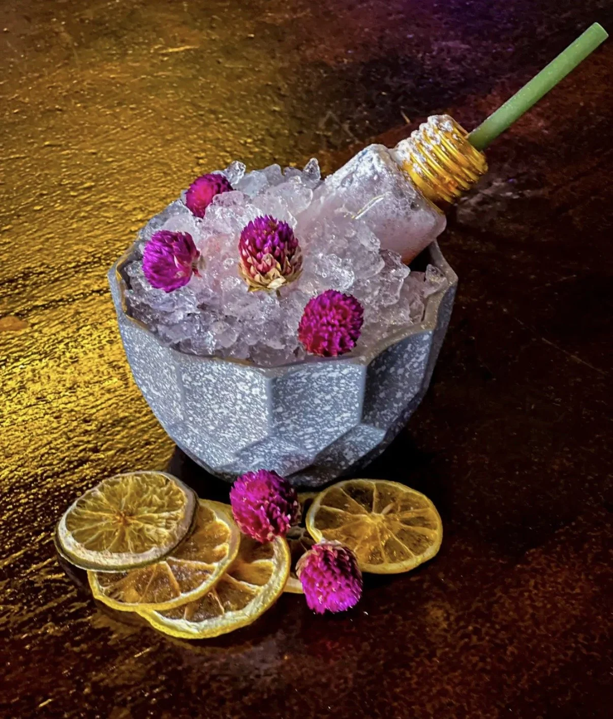 A glass bowl filled with ice and decorated with pink flowers, with a small bottle inserted like a straw, sitting on a dark wooden surface with sliced dried lemon pieces nearby.