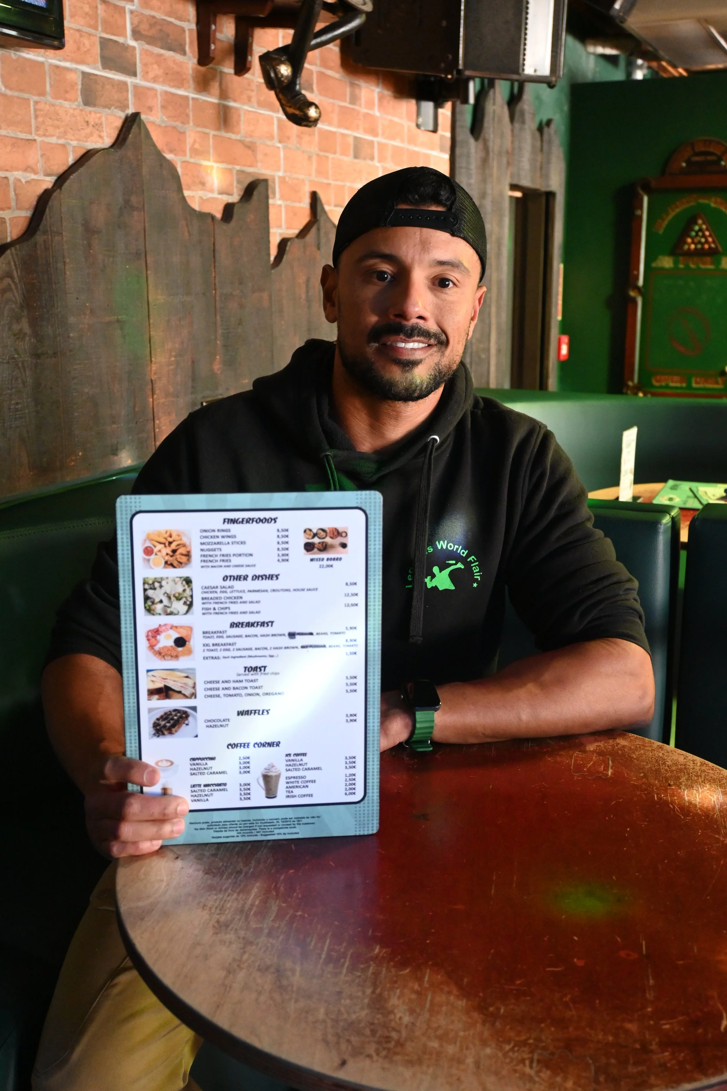 A man sitting at a wooden table in a restaurant, holding a menu. He is wearing a black hoodie, a black baseball cap worn backwards, and a smartwatch. The background features a brick wall, green decor, and wooden accents.